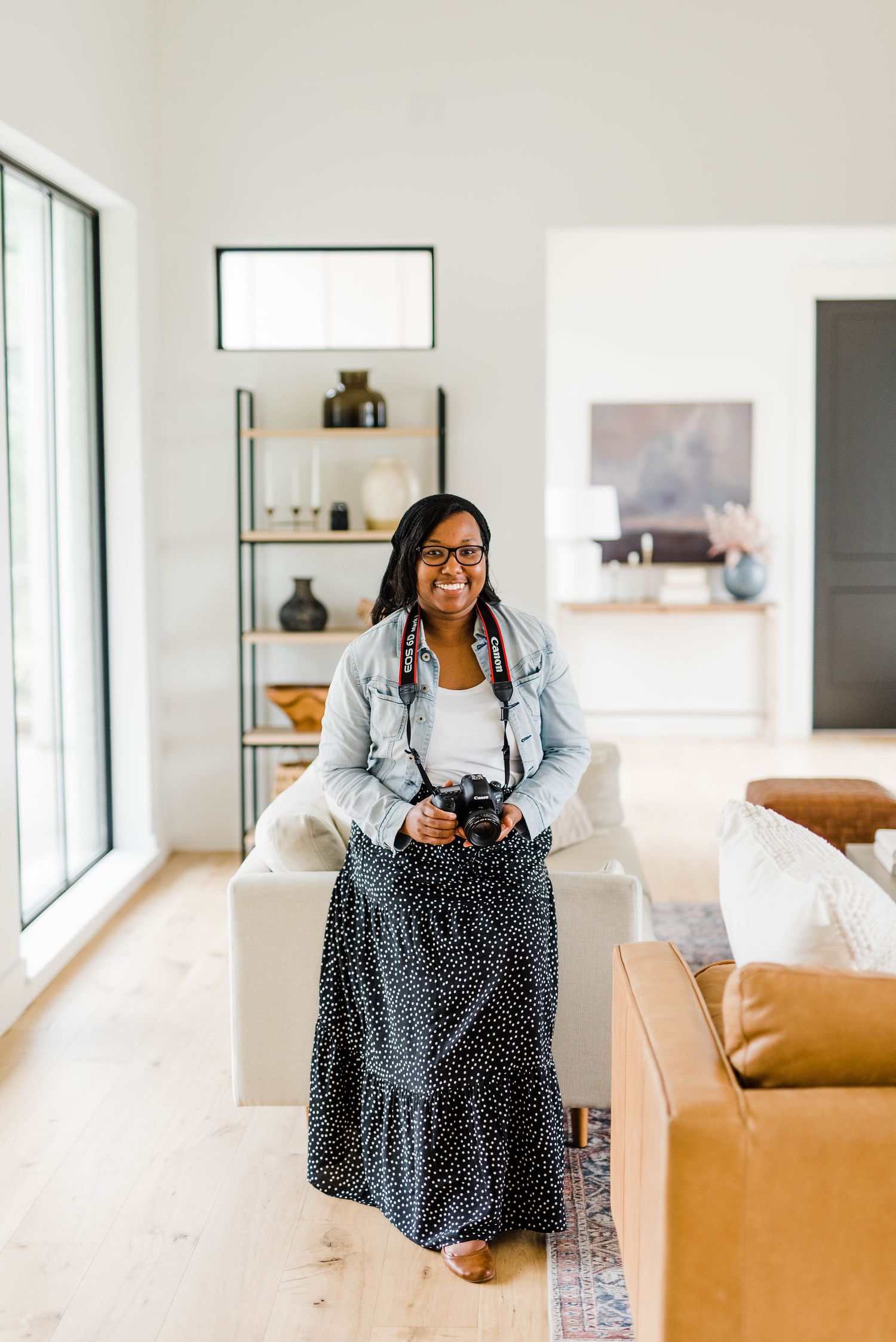 Woman in denim jacket and polka dot dress sits casually on modern couch in bright living room interior.