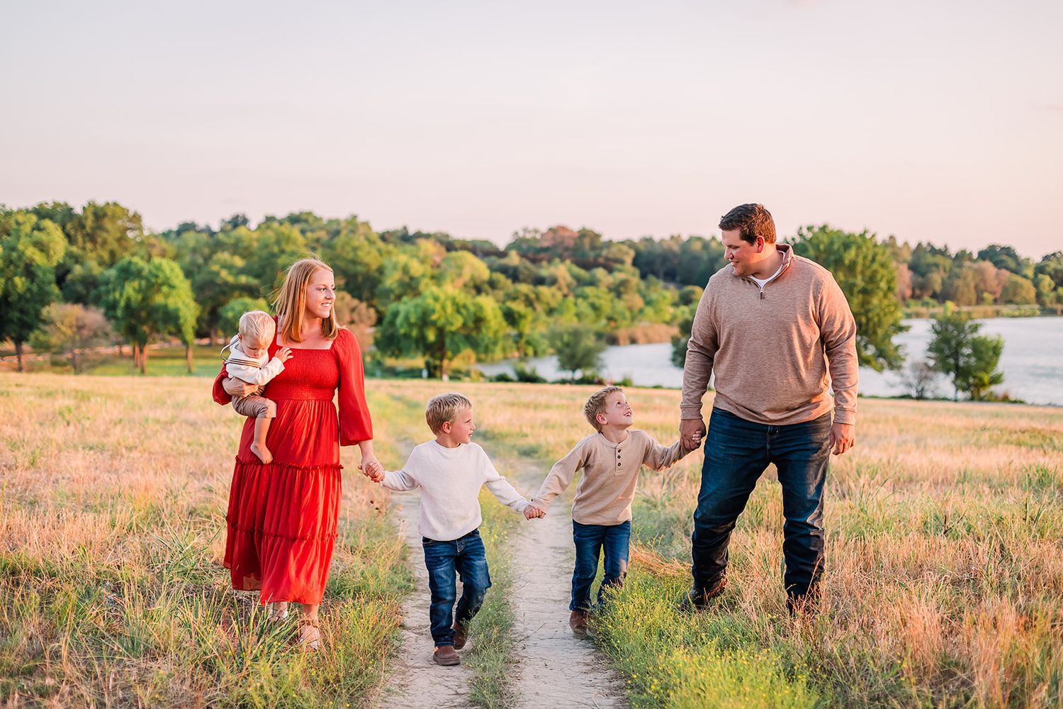 Family walking together on a trail through a golden meadow at sunset near a lake.