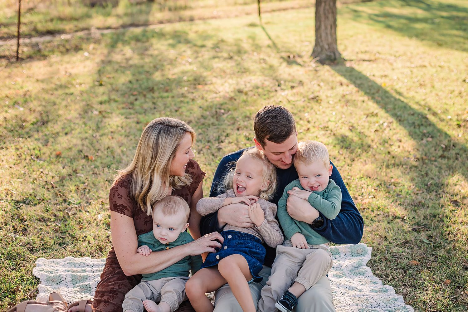A family cuddles together on a blanket in a sunny park setting.