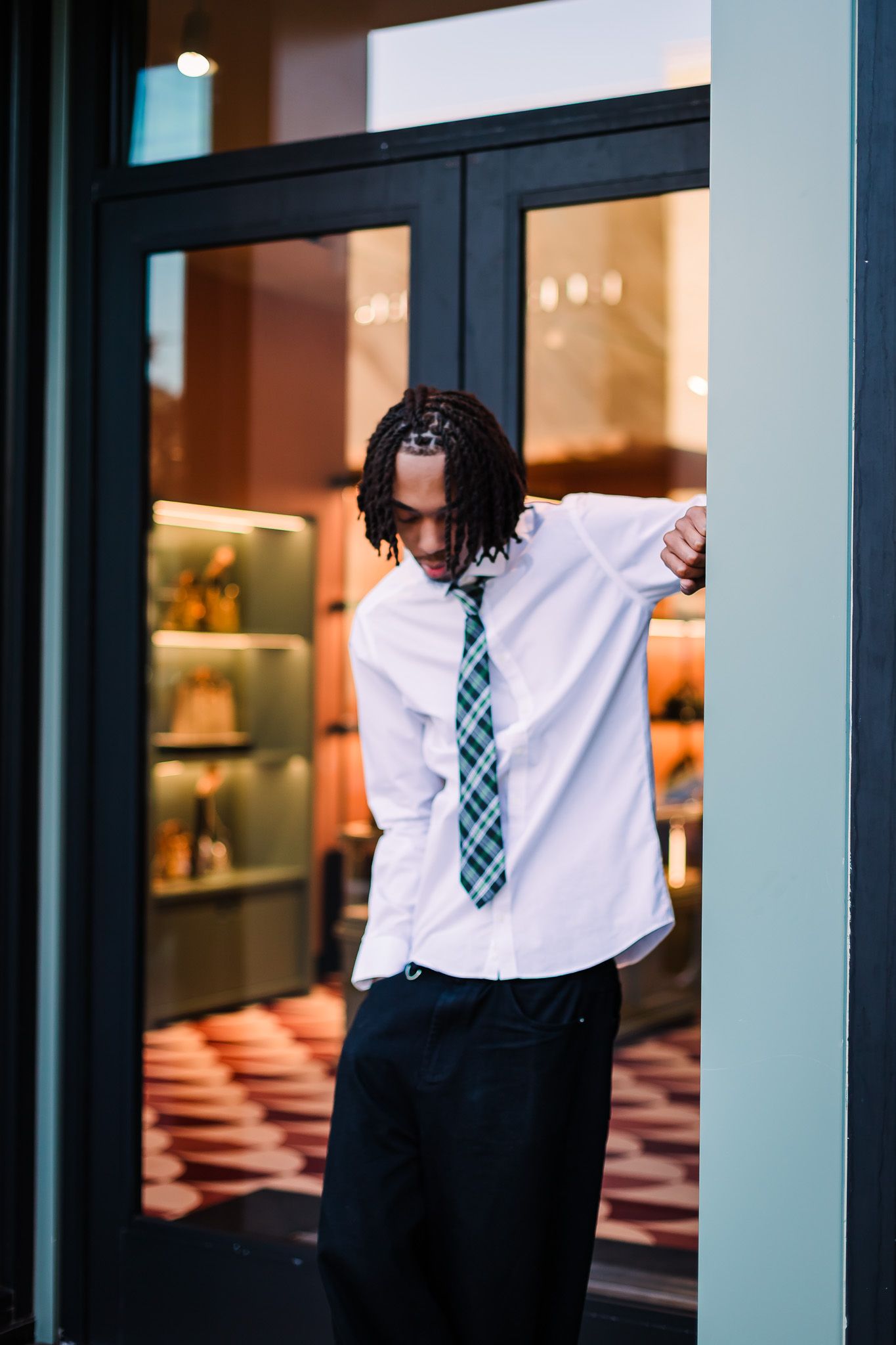 A person in business attire with a tie poses expressively against a glass window in an urban setting.