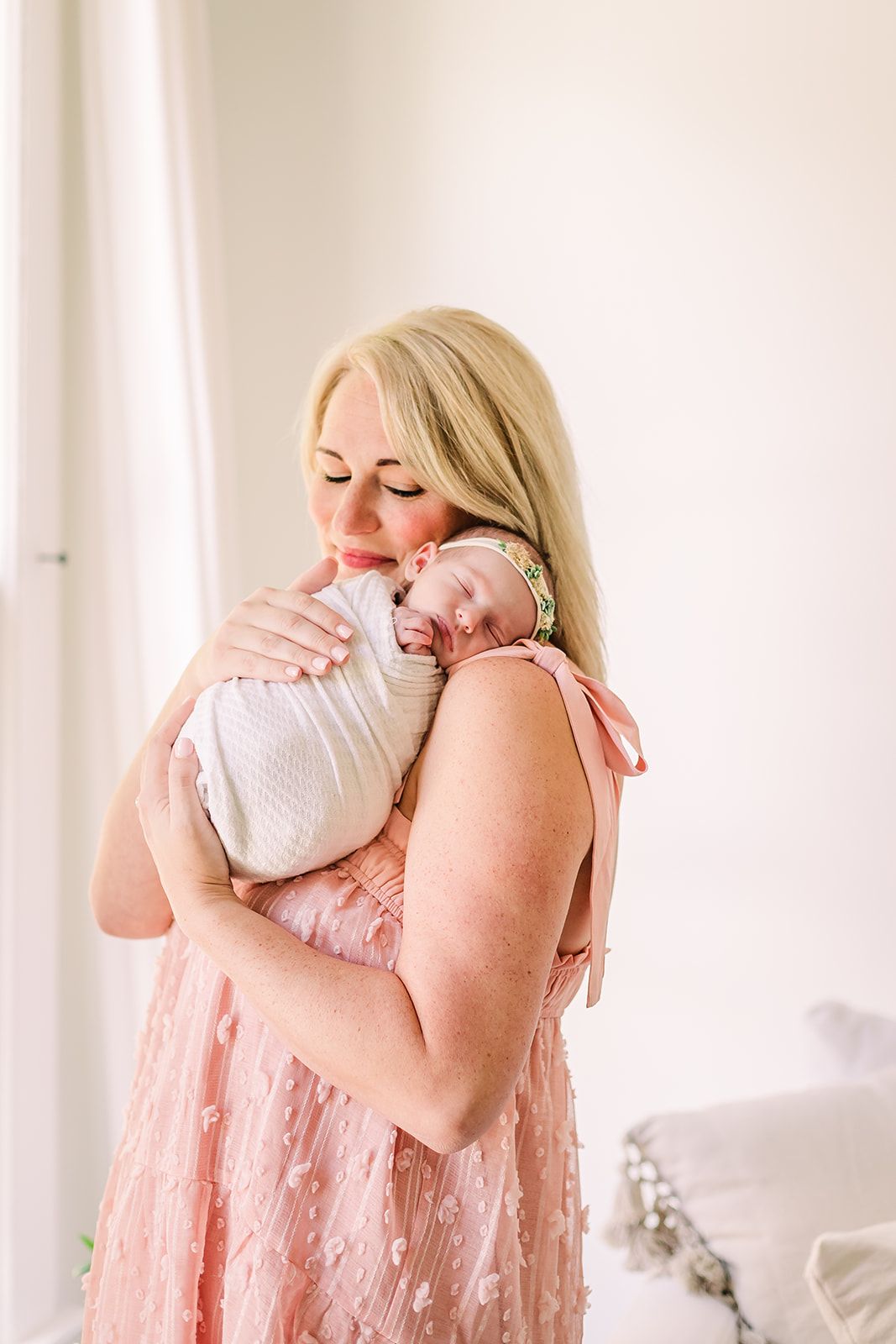 A tender moment captured in soft lighting as a parent embraces a baby in a white blanket while wearing a pink dress in McKinney TX