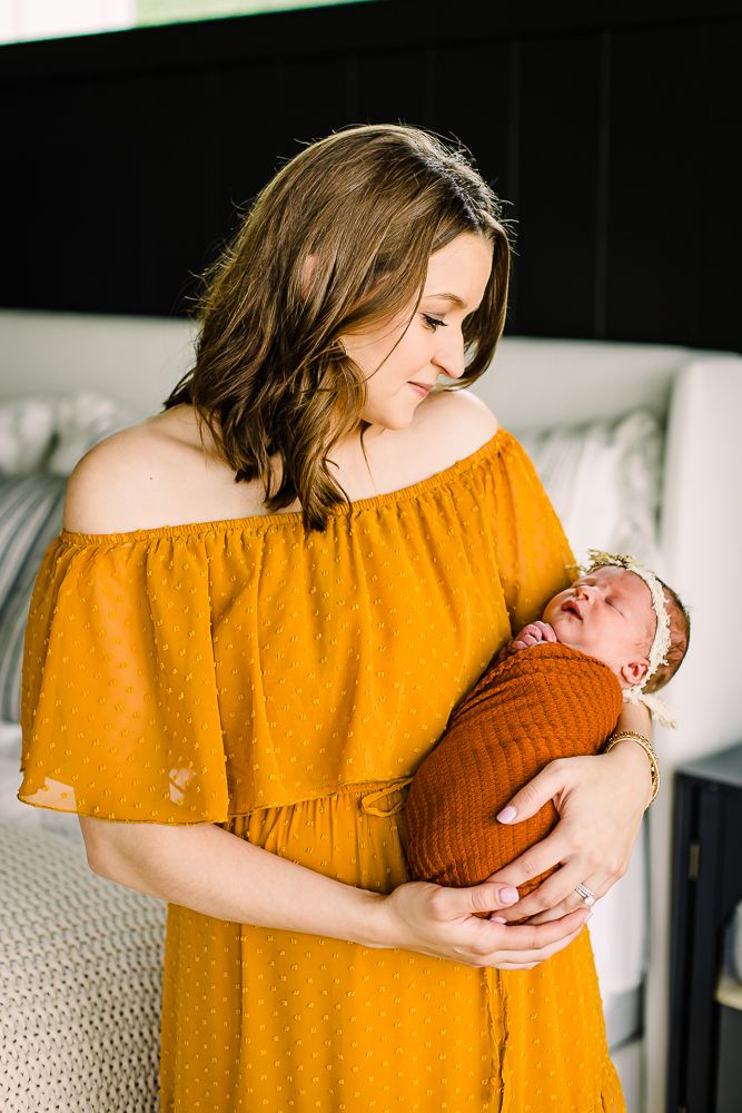Mother in mustard yellow dress cradles sleeping newborn baby in matching knit outfit on white bedding.