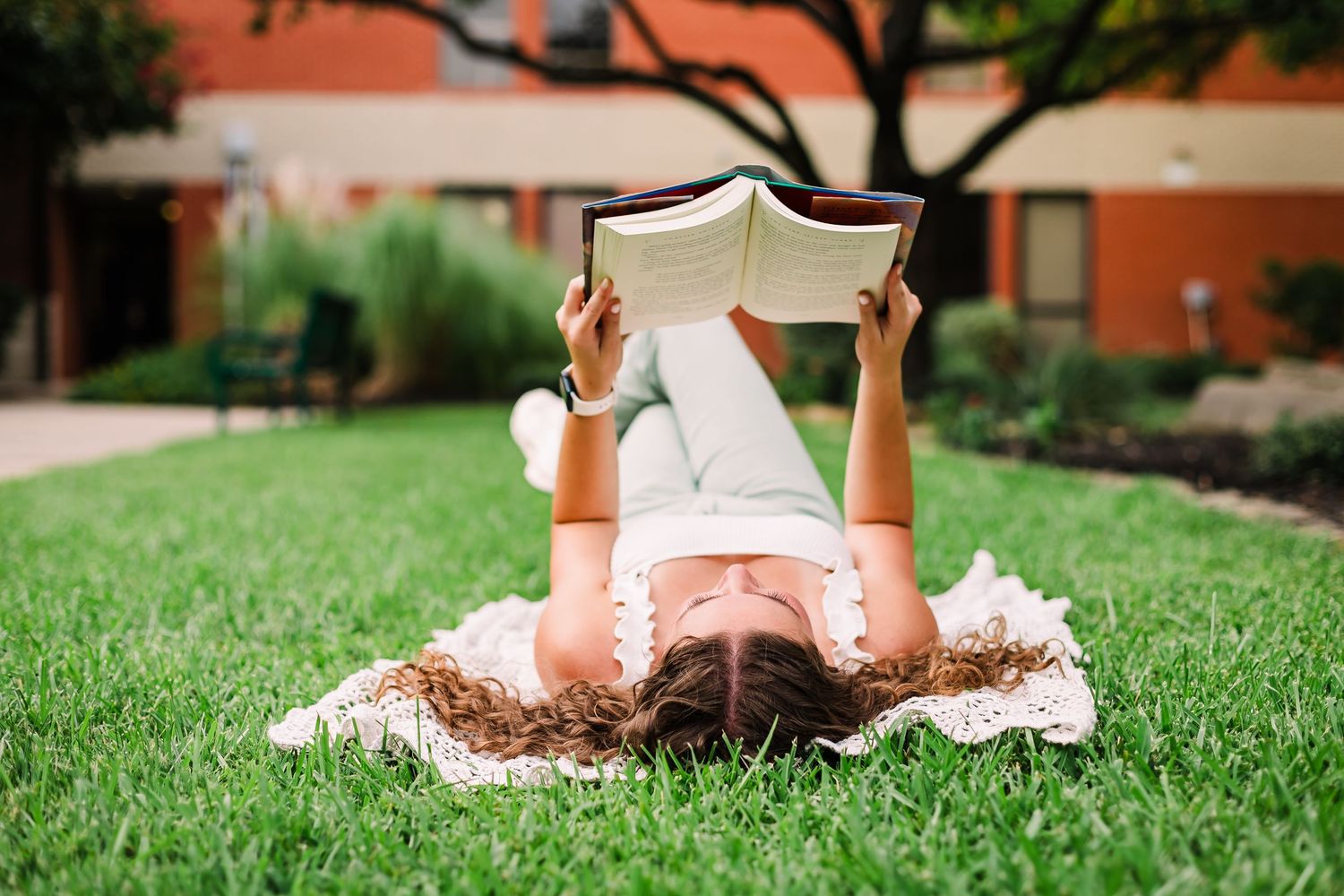 Person lying on grass reading book during sunny day outdoors.