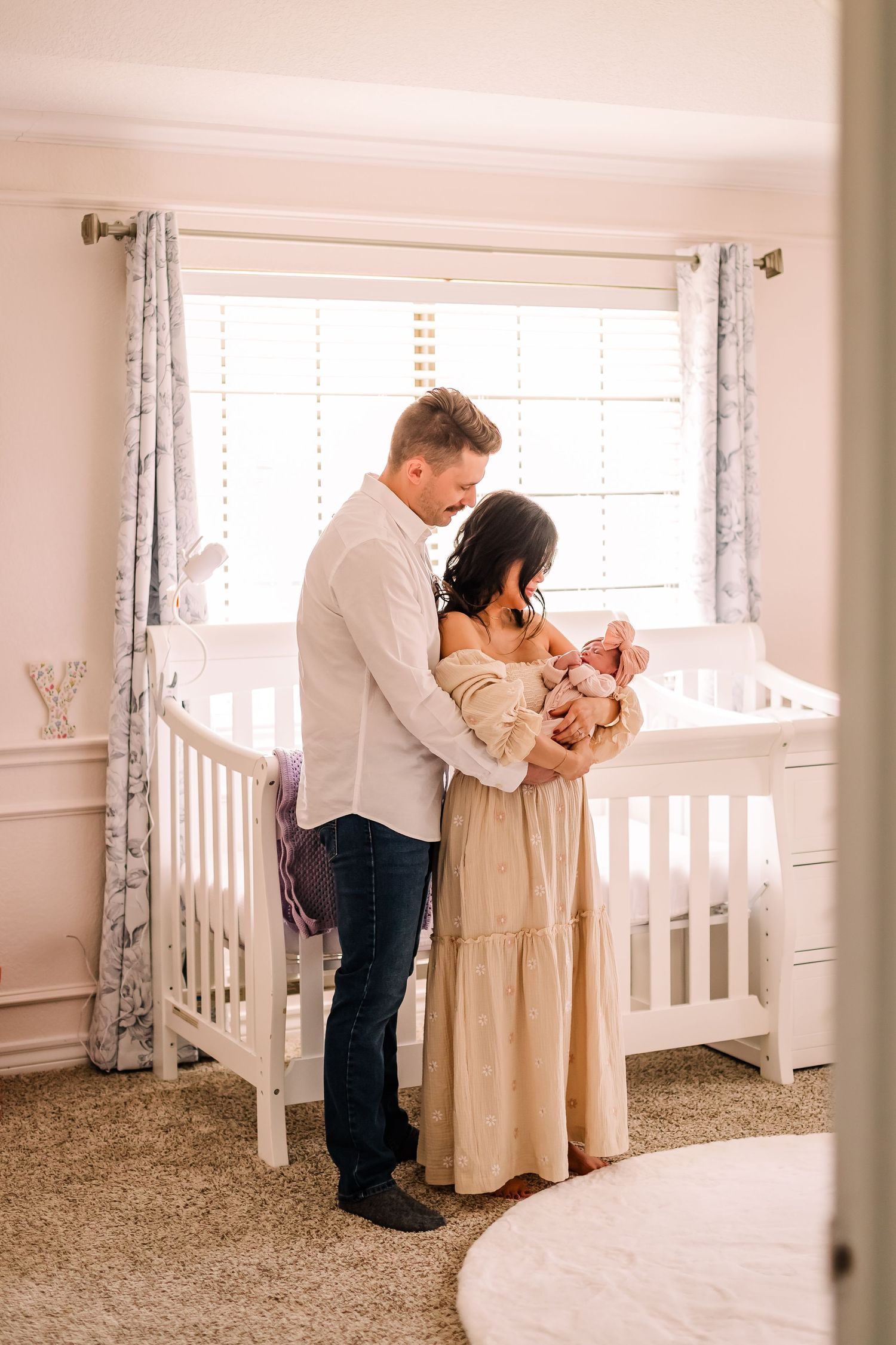 Parents lovingly embrace their newborn baby next to a white crib in a bright nursery room.