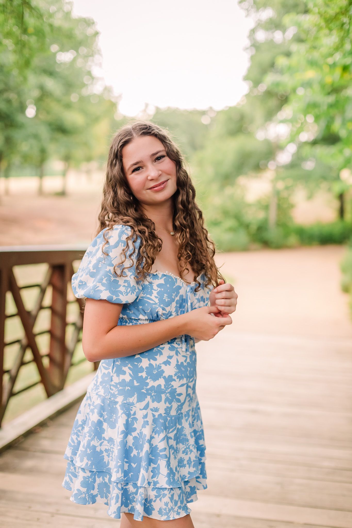 Person in a blue floral dress poses on a wooden bridge in a natural setting.