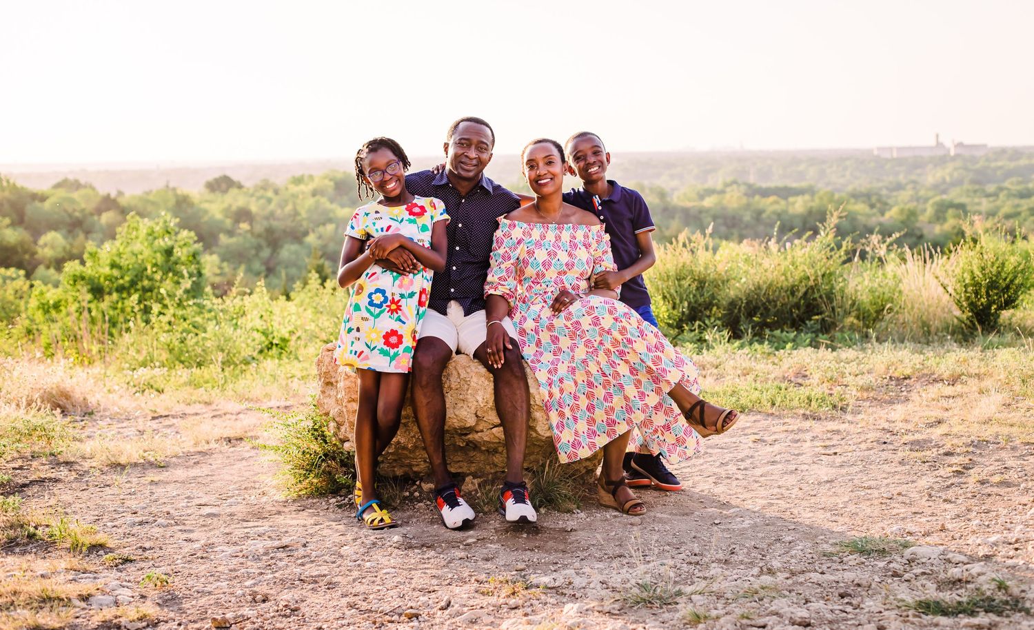A group of people in floral dresses sit together on a bench in a rustic outdoor setting at sunset.