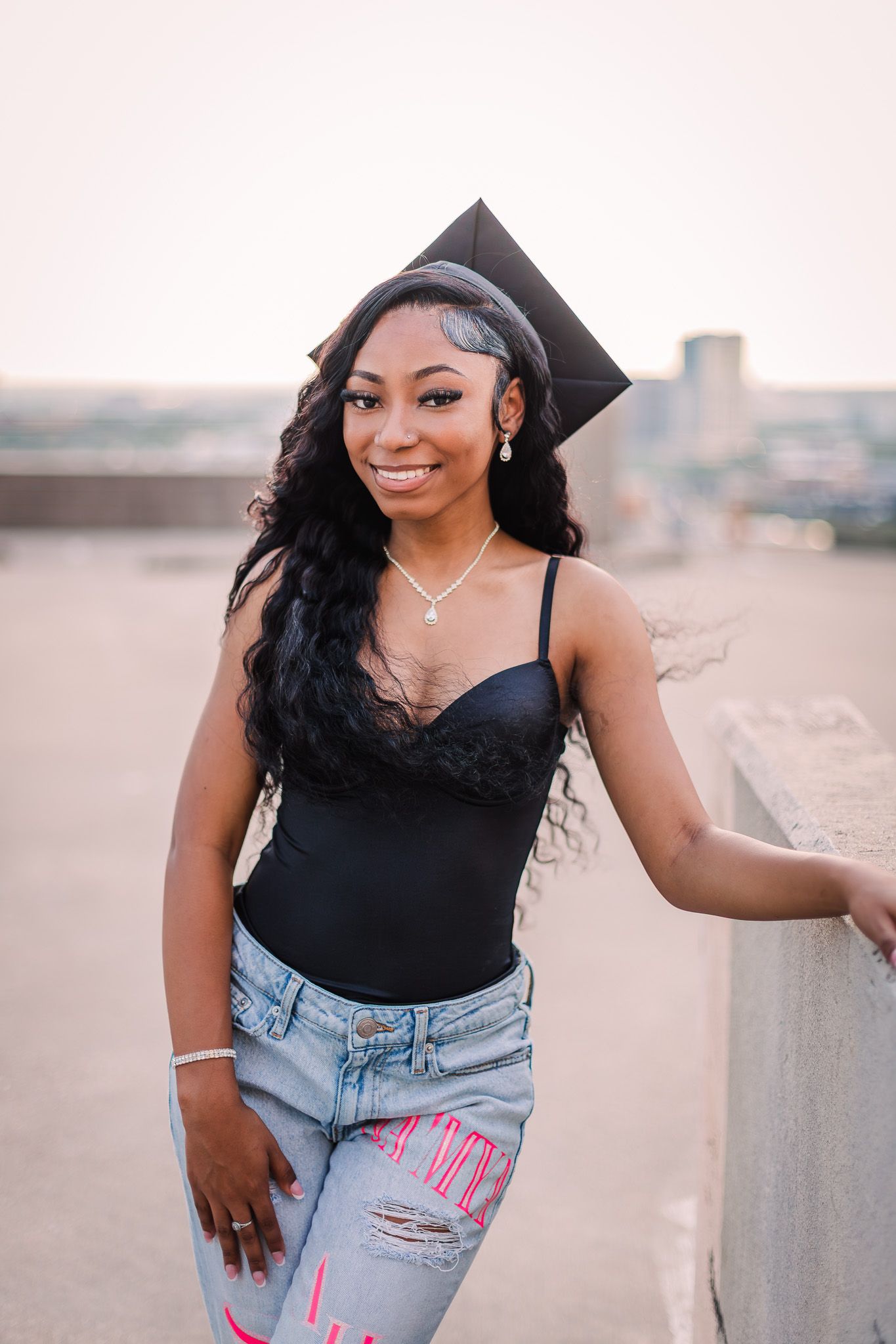 A graduate in a black top and denim poses outdoors during a celebratory photoshoot at sunset.