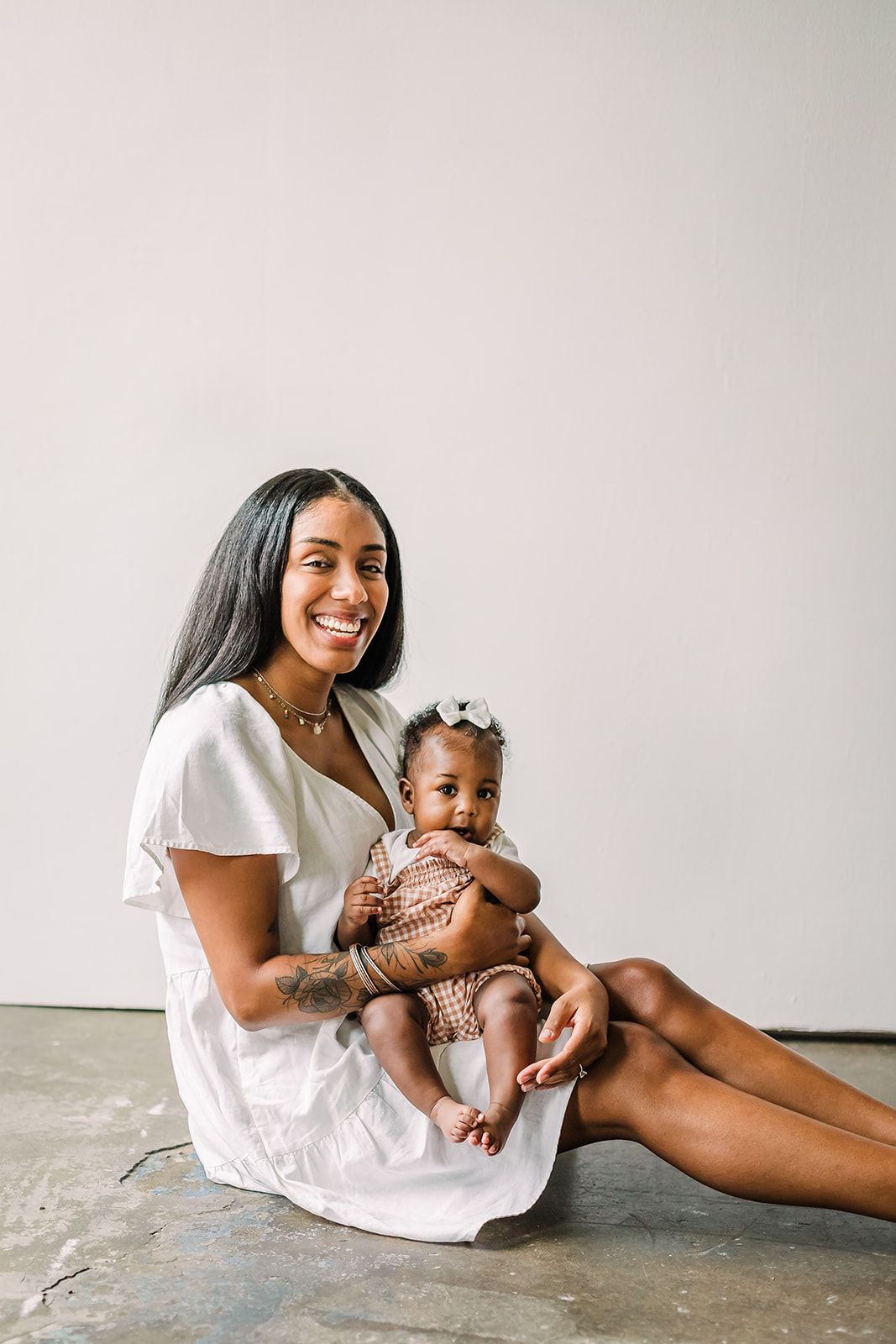 A mother and baby share joyful moments together while sitting on a light colored floor in matching white outfits.