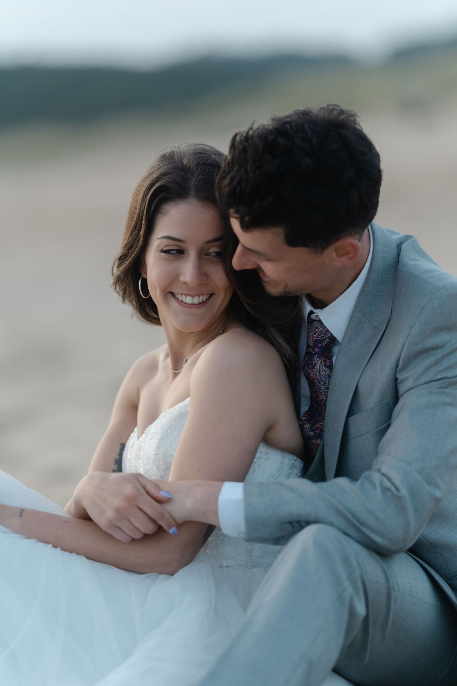 Smiling bride in white dress embraced by groom in gray suit on beach during golden hour.