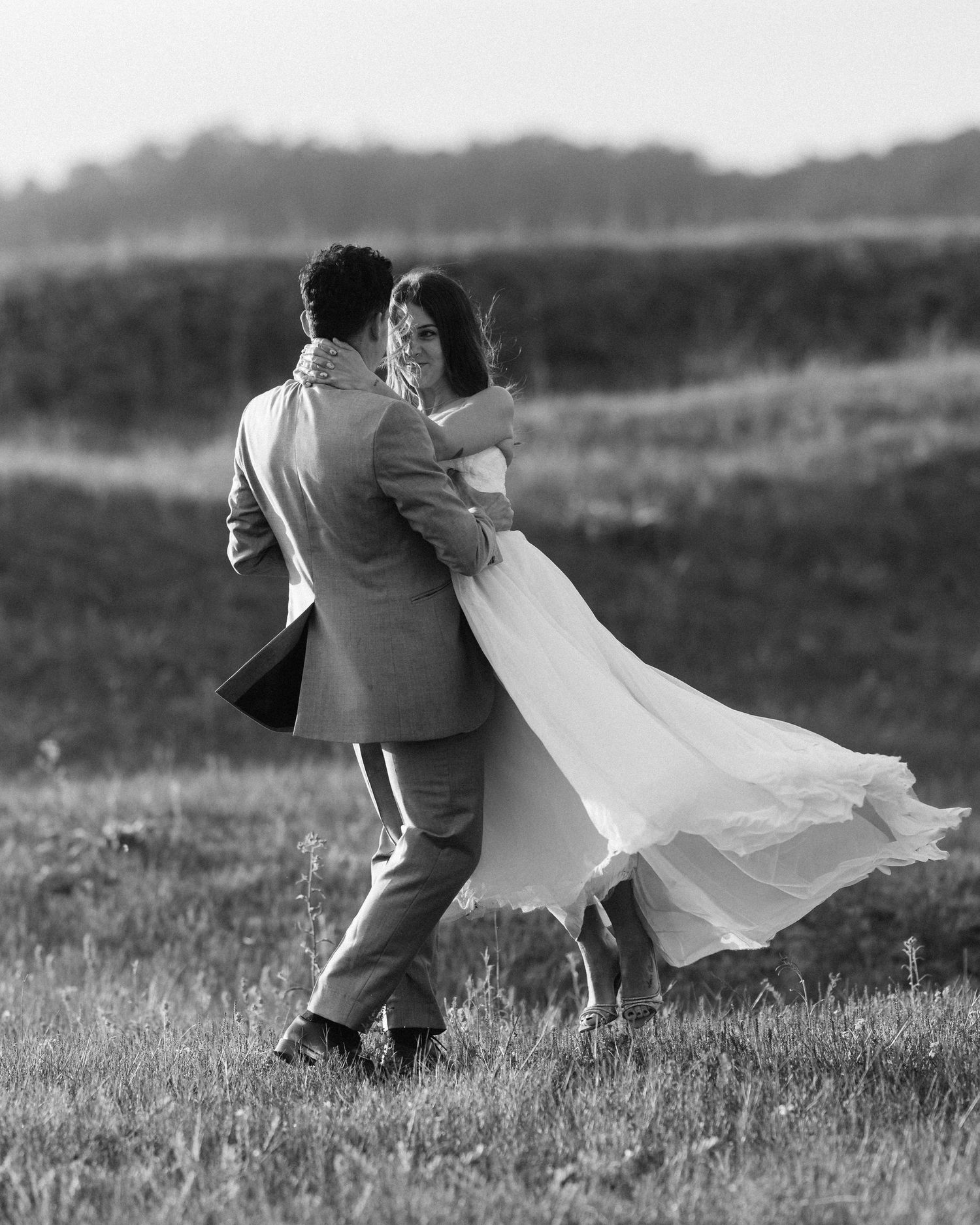 Groom lifts bride in a joyful spin in a sunlit field in black and white wedding photo.