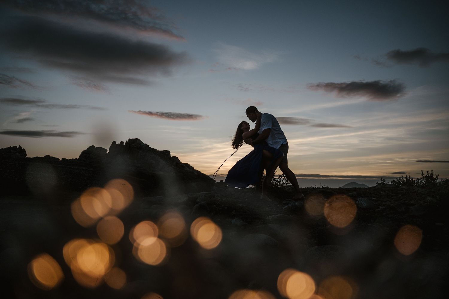Silhouettes of figures against a dramatic sunset sky with glowing orange bokeh lights in the foreground.