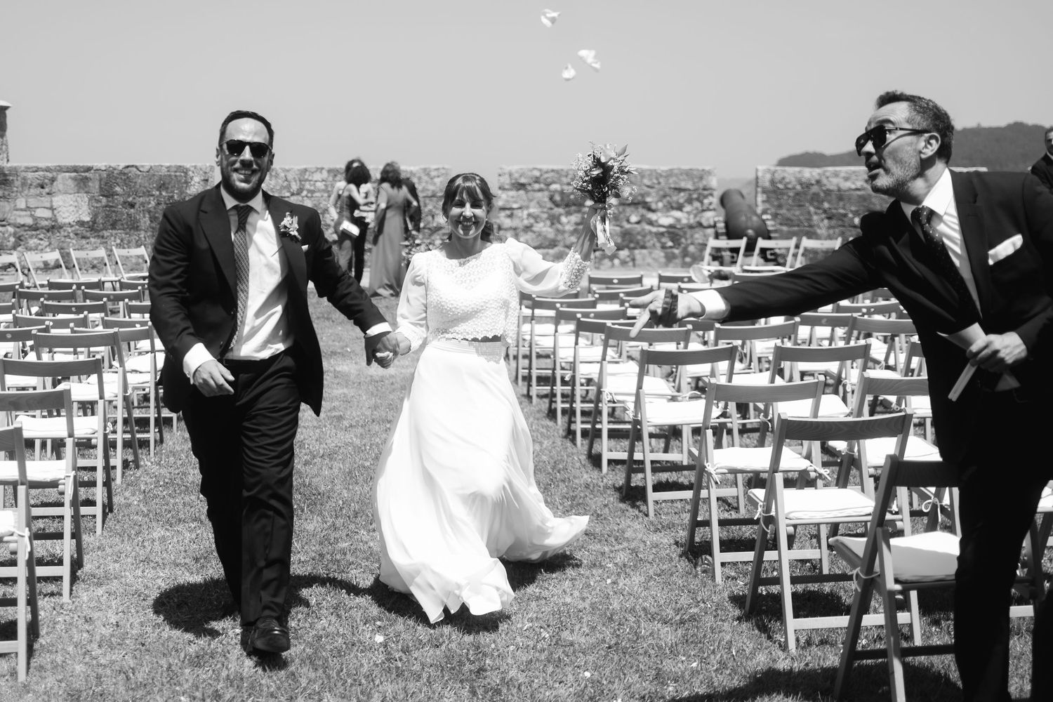 Wedding party members playfully pose and jump in front of white ceremony chairs on a lawn.