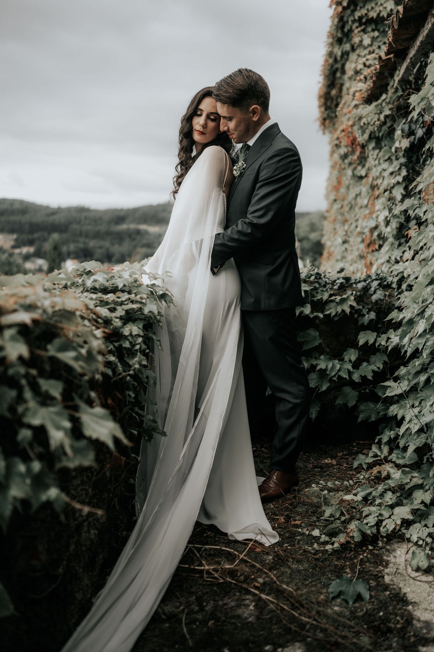 A romantic wedding couple embraces near rocky cliffs in a moody, dramatic outdoor setting.
