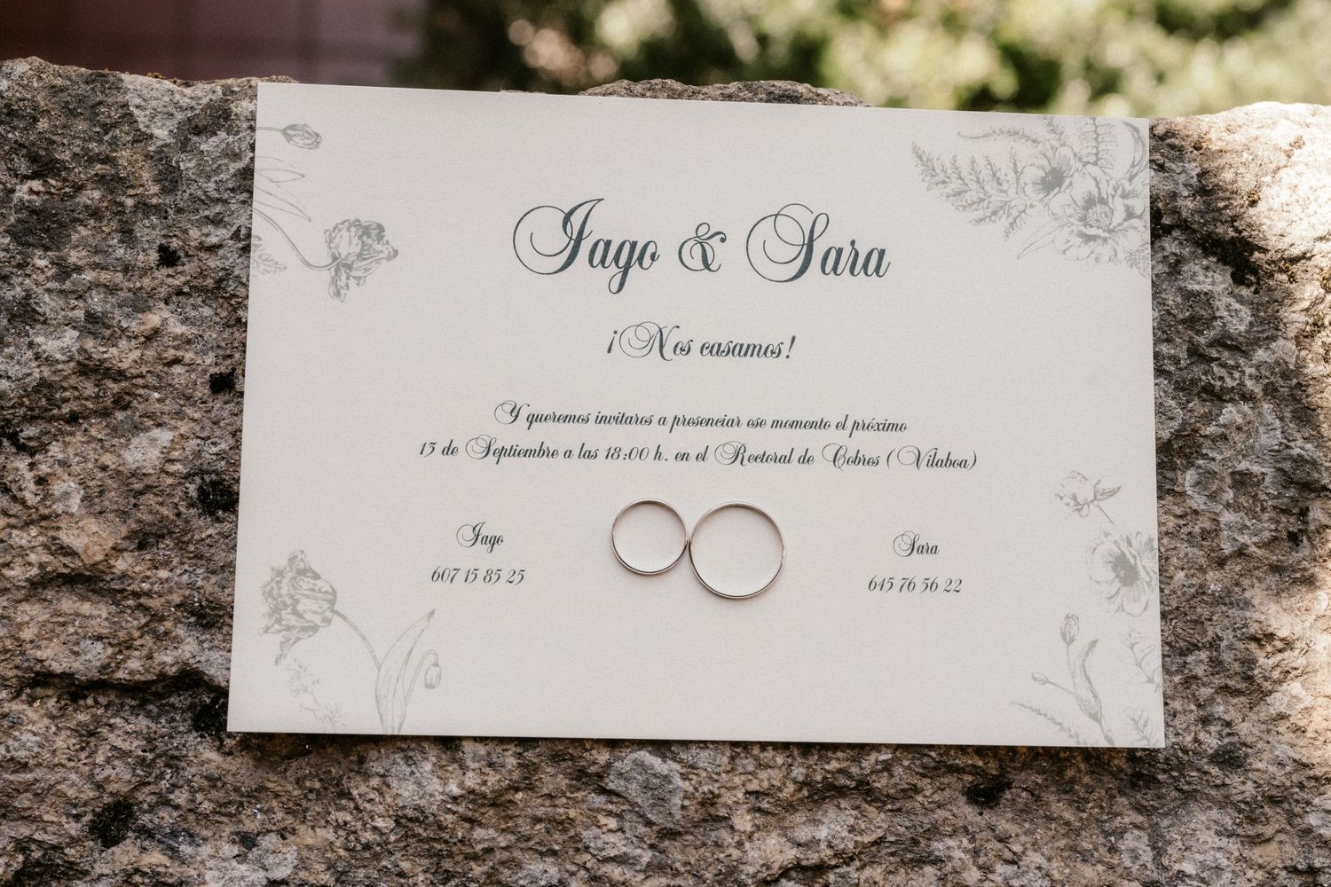 Wedding invitation card with two gold wedding rings placed on stone surface with delicate floral design details.