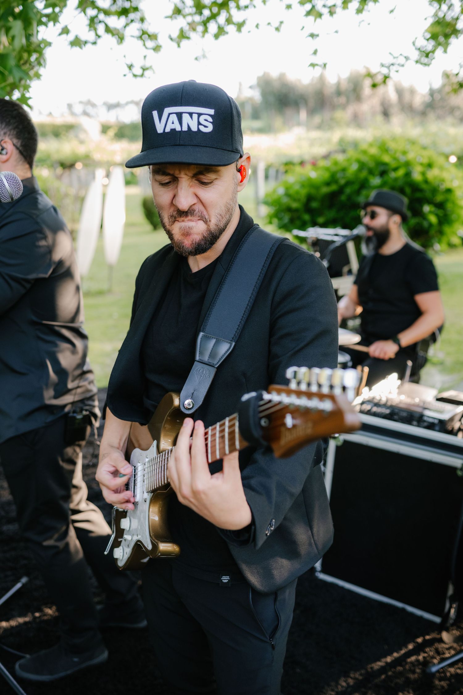 Musicians in black attire perform with guitars at an outdoor event venue.