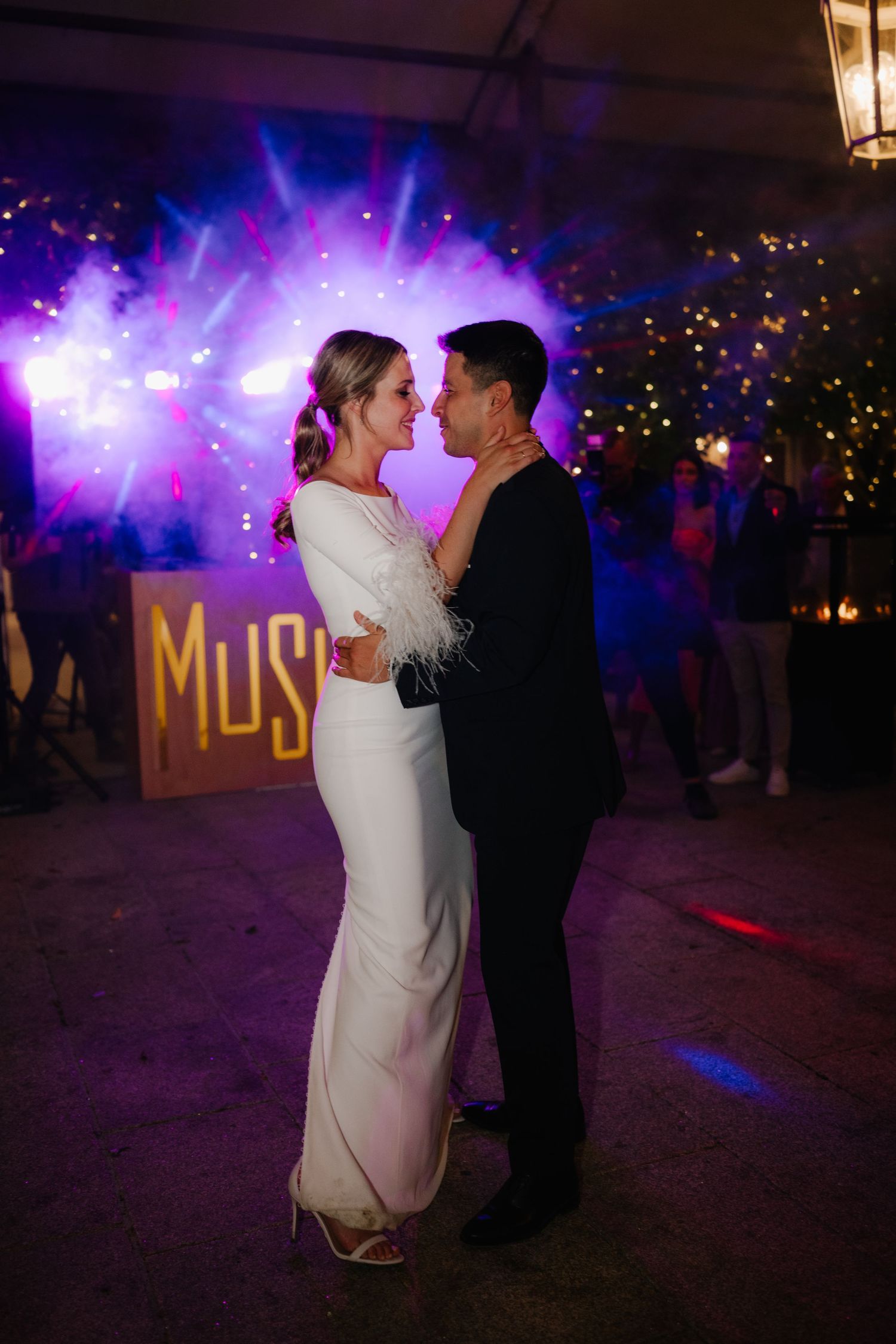 Couple shares a romantic dance under purple lighting with illuminated MUSIC sign in the background at evening reception.