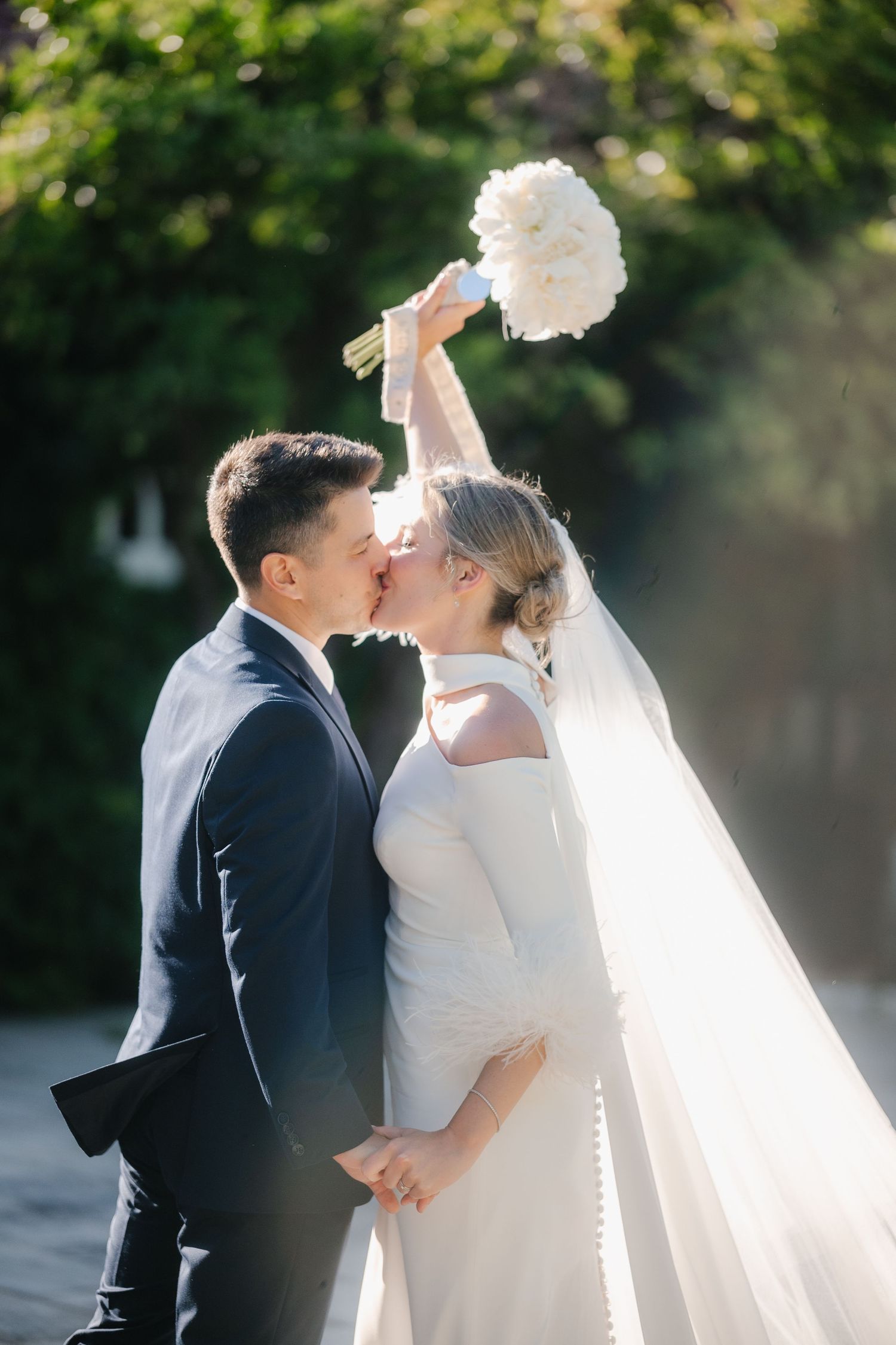 A romantic wedding couple shares tender moments in a sunlit garden setting with flowing white veil and formal attire.