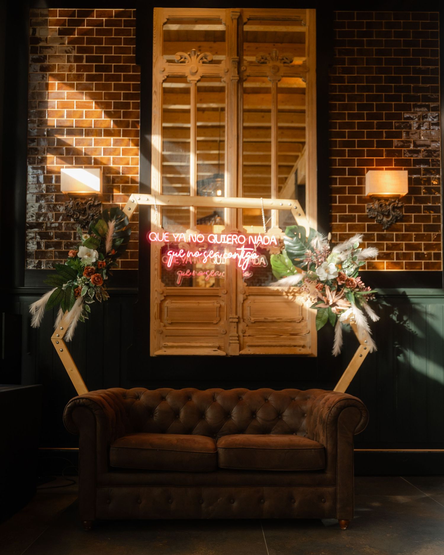 Brown leather chesterfield sofa beneath wooden shutters and neon sign on exposed brick wall with ambient lighting.