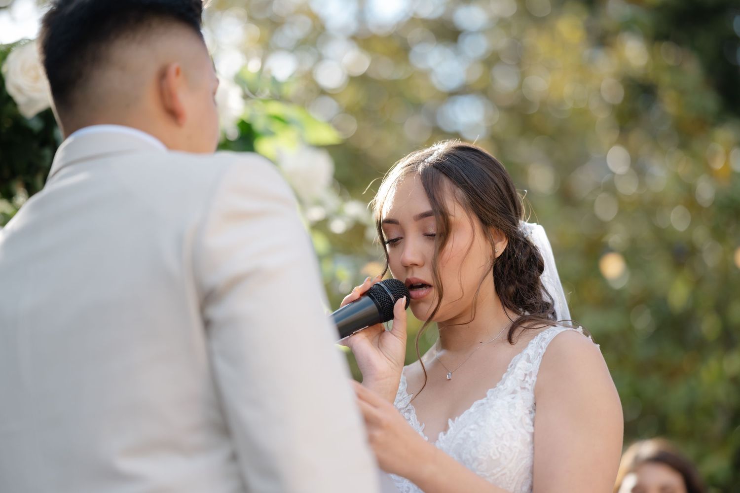 A bride in a white lace dress reads her wedding vows while holding a microphone during an outdoor ceremony.