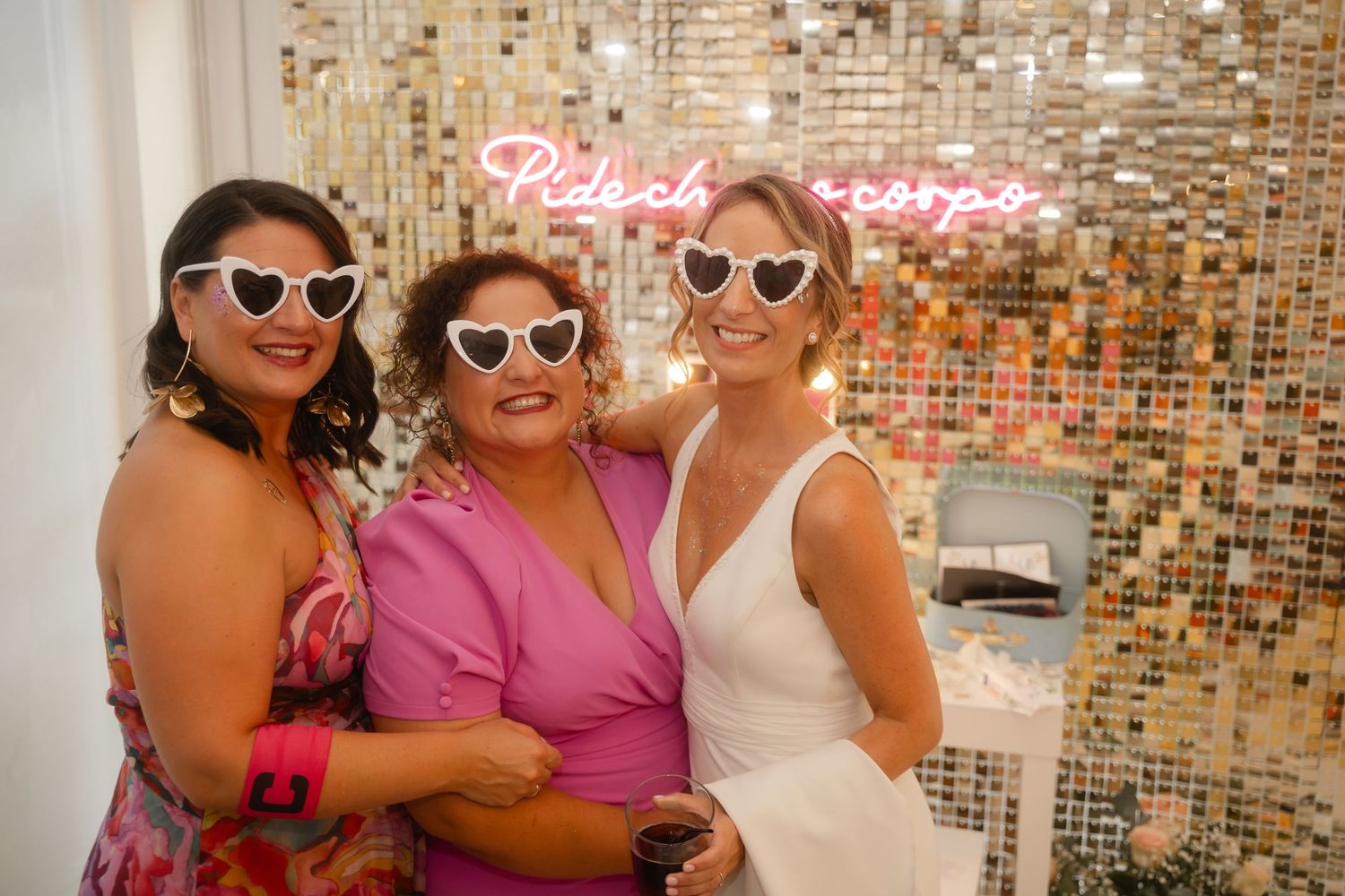 Three people wearing sunglasses pose together against a sparkly gold sequin wall with pink neon signage.