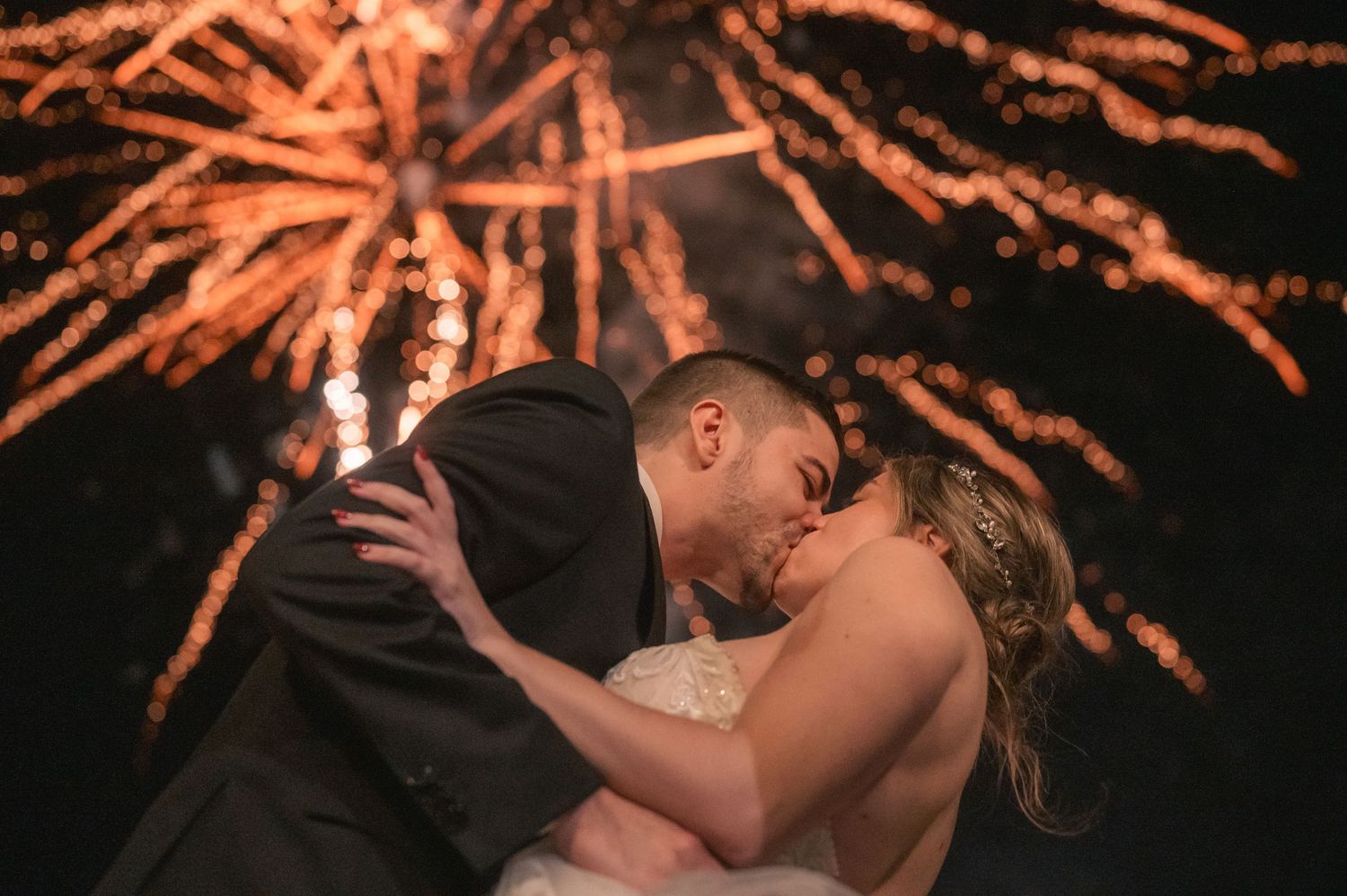 A couple shares a romantic kiss under warm fireworks lighting up the night sky during a celebration.