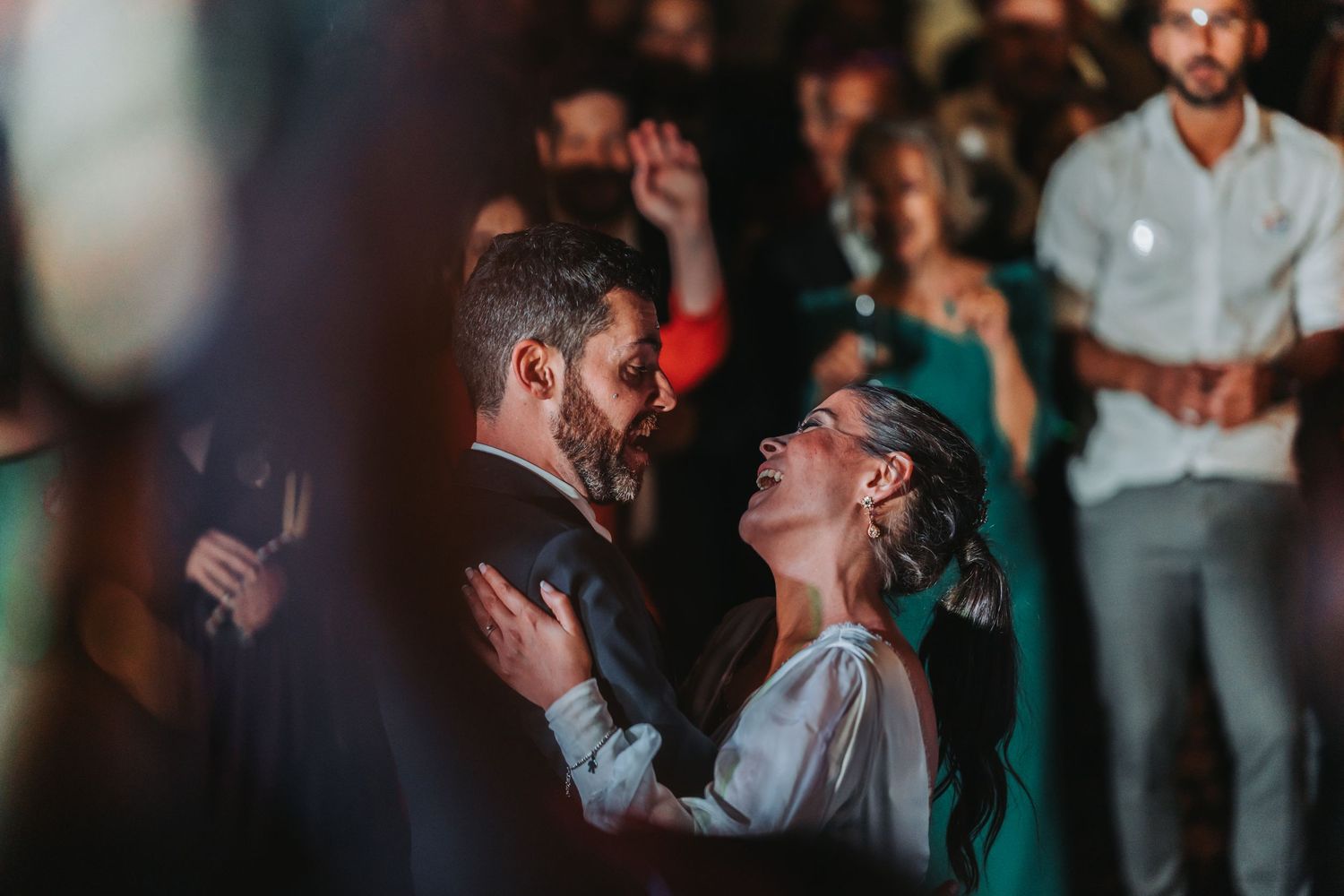 A couple shares an intimate dance moment during a wedding reception with atmospheric lighting and guests in the background.