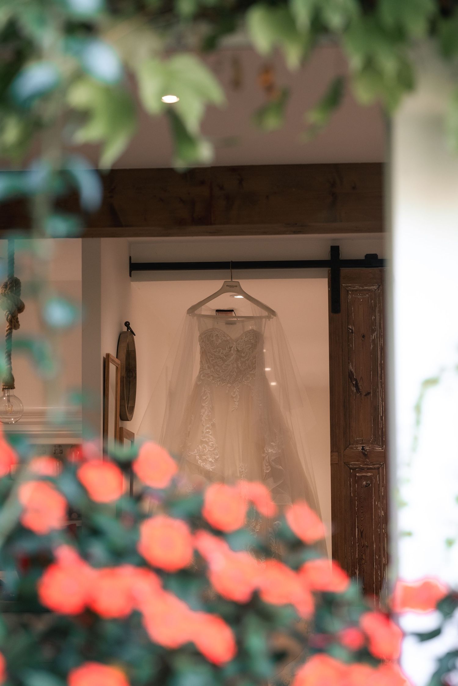 A delicate white lace wedding dress hangs in a doorway framed by coral-colored flowers in soft focus.