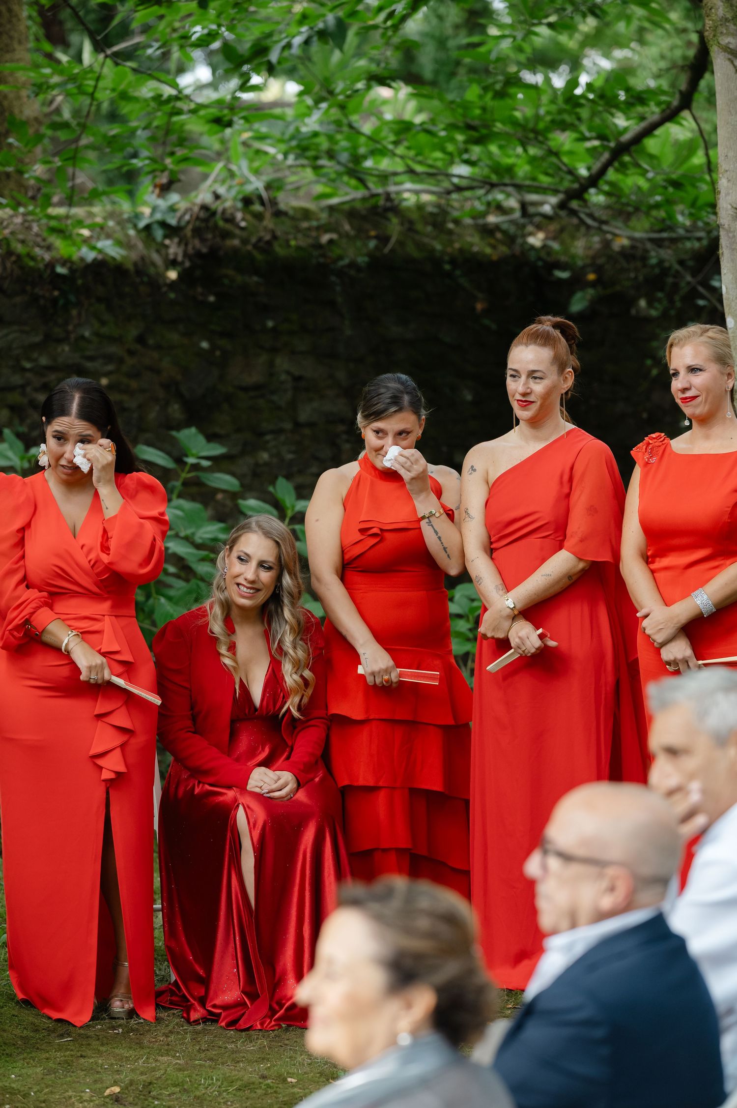 A row of bridesmaids in matching red dresses stand together during an outdoor wedding ceremony.