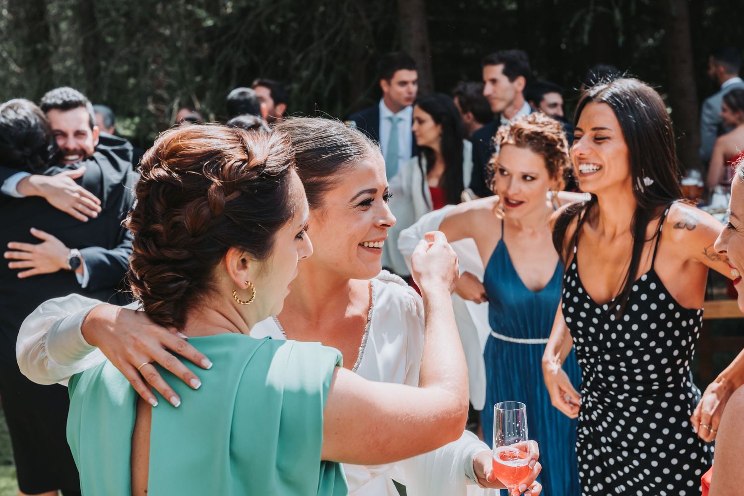 A group of wedding guests laugh and celebrate together while holding drinks at an outdoor reception.