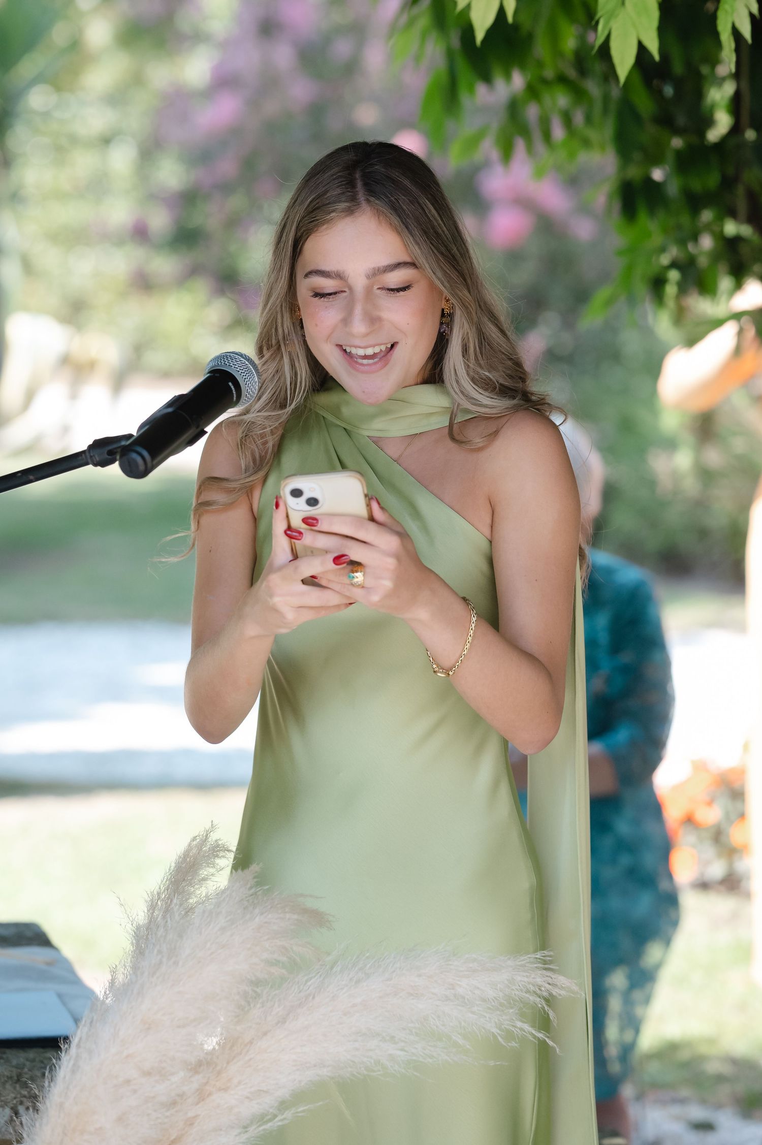 Someone in a light green dress reads from a phone while standing at an outdoor podium during a sunny day.