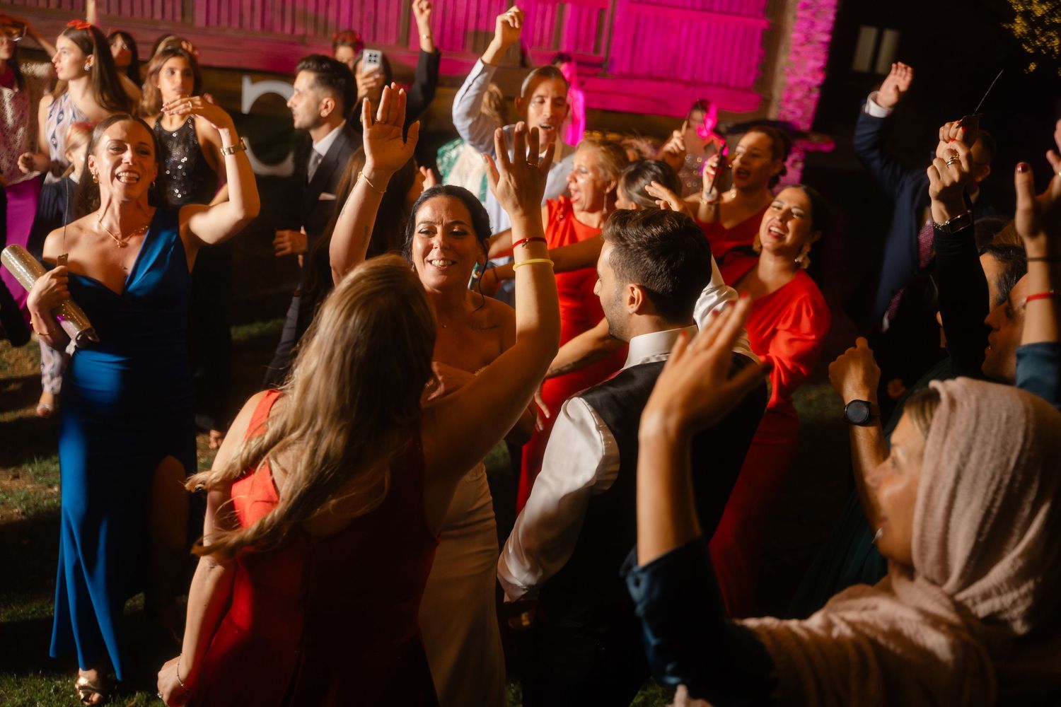A large group of people dance and celebrate at a wedding reception with colorful pink lighting in the background.