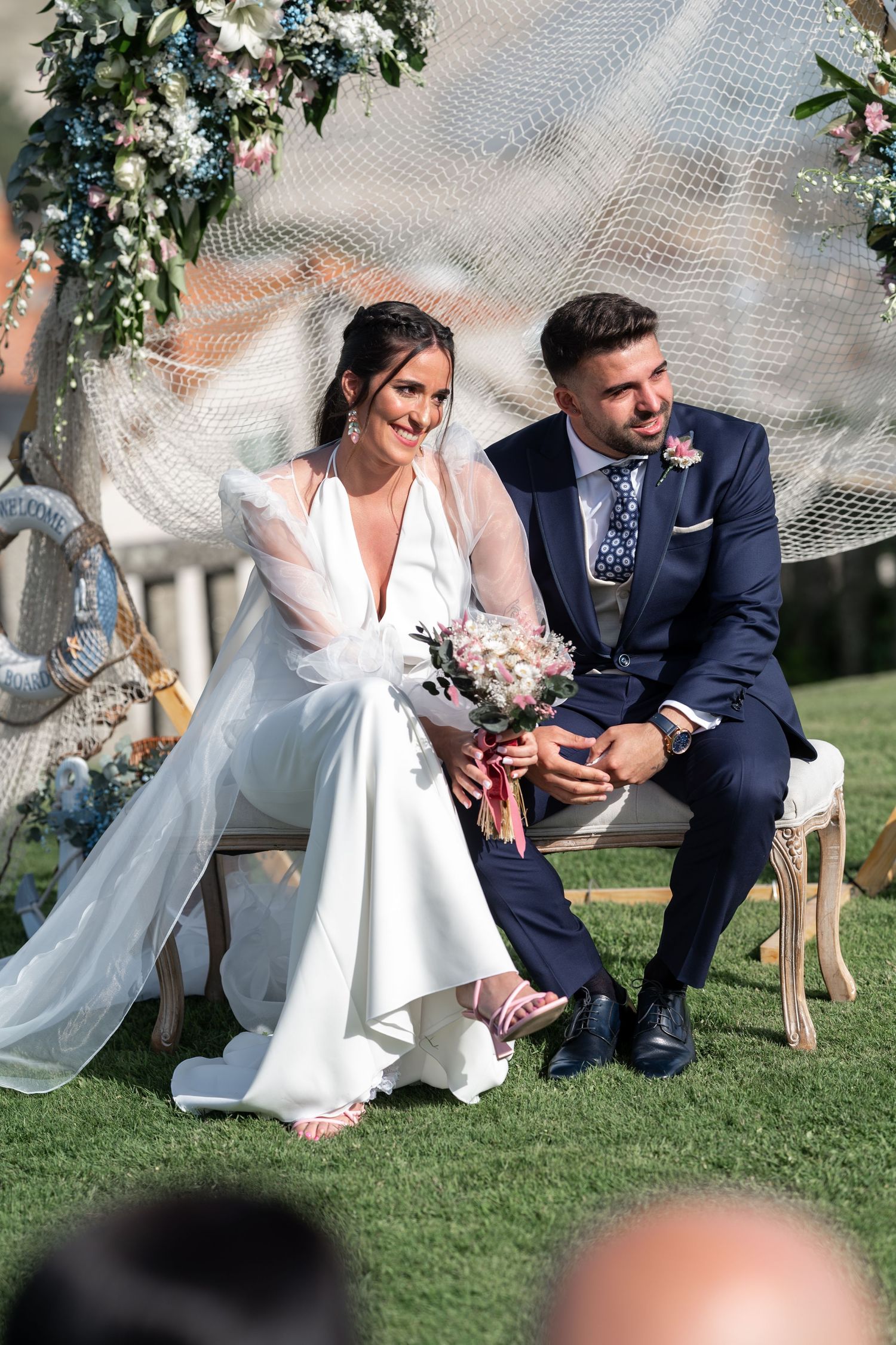 Wedding couple seated on grass under floral arch during intimate outdoor ceremony.