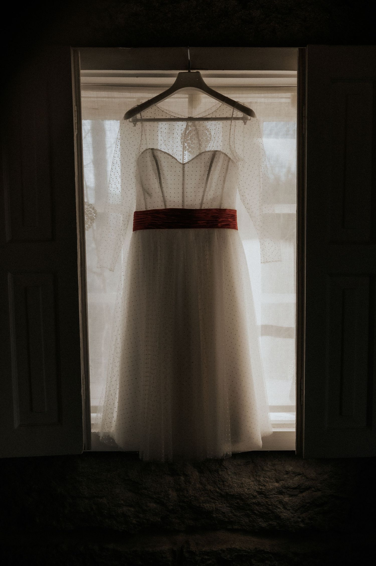 A white wedding dress with a red sash hangs in silhouette against a bright window in multiple artistic views.