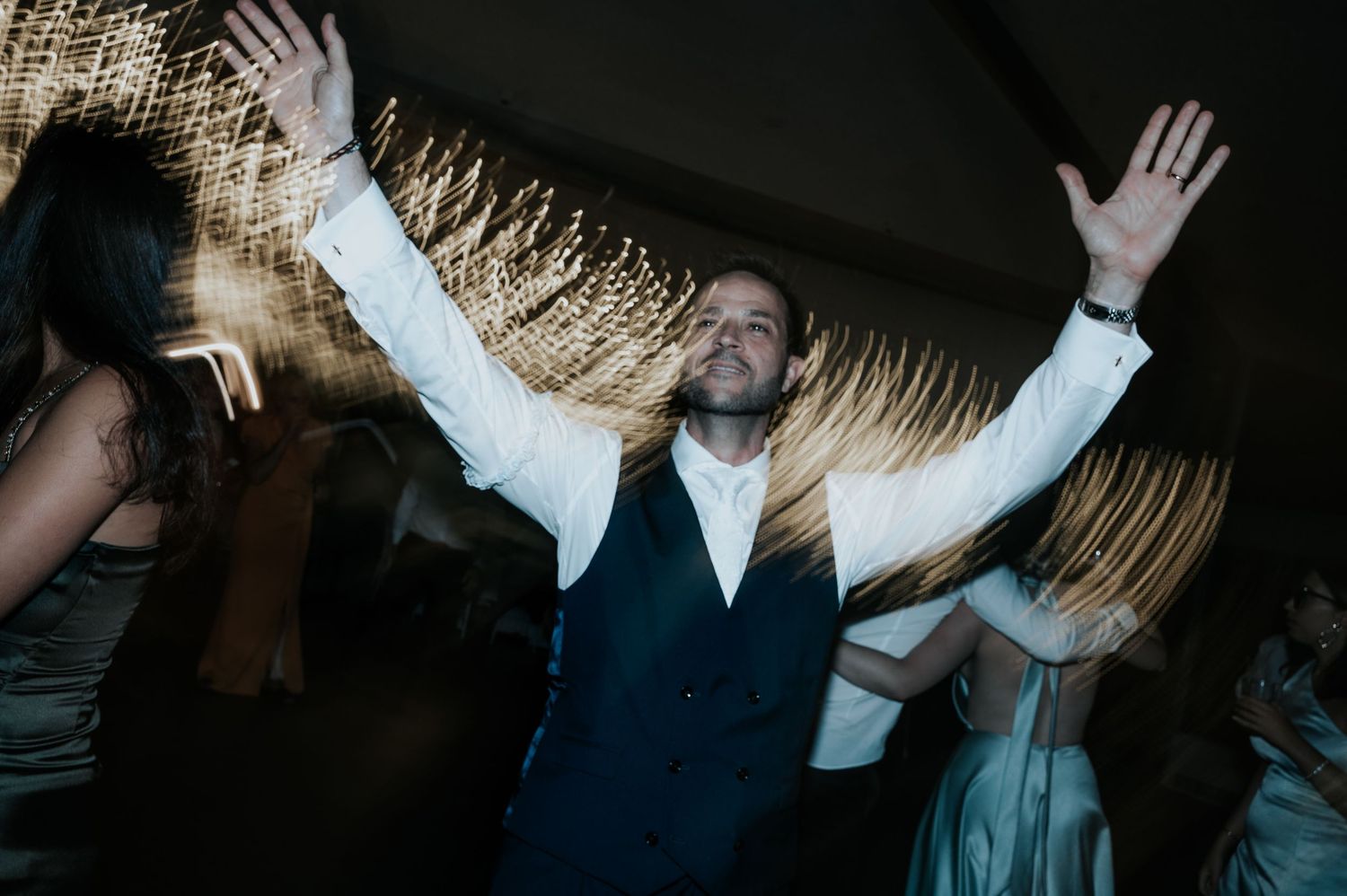A group of people dancing with raised hands in a dark room with light trails and motion blur effects.