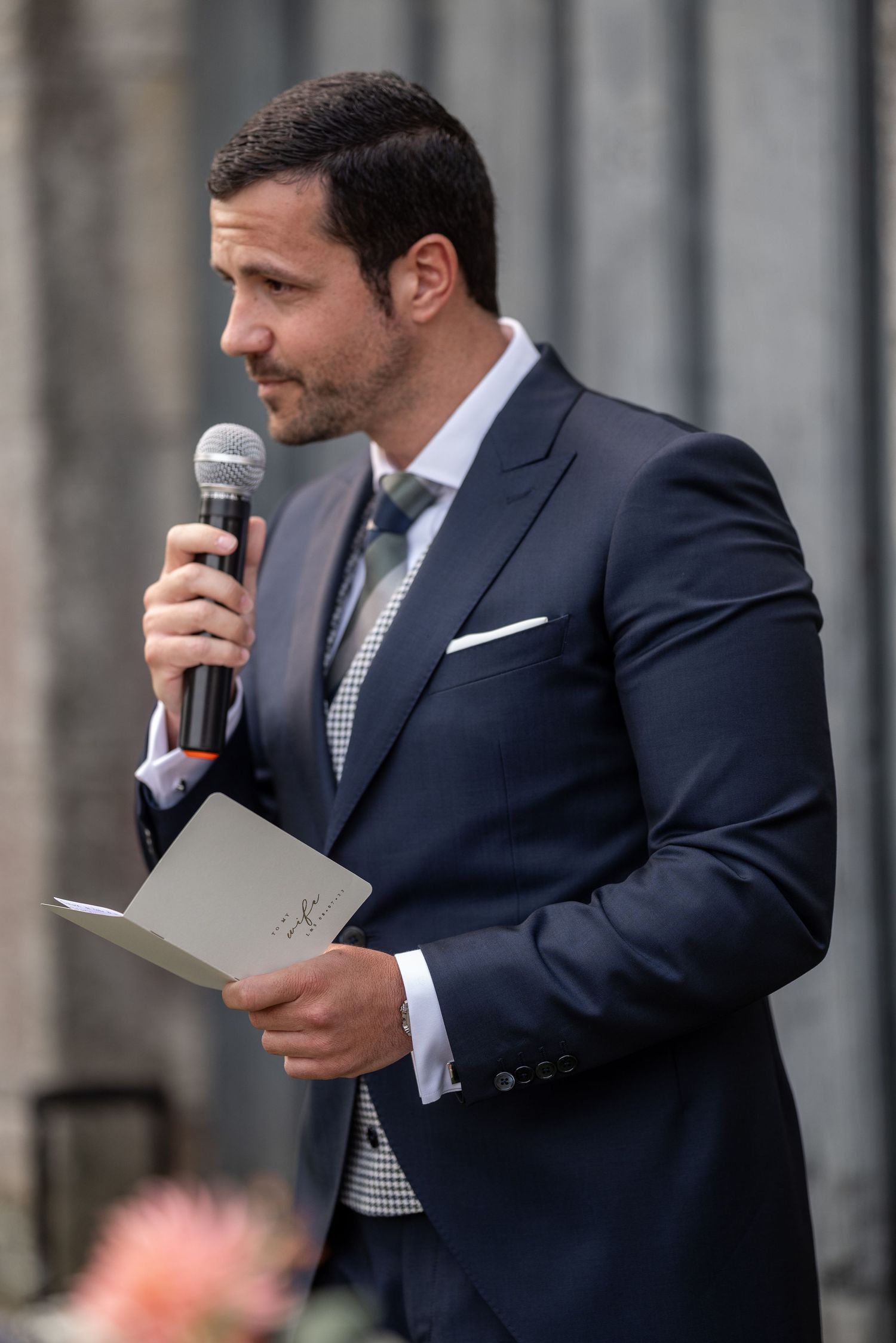 A person in a dark suit delivers a wedding speech while holding a microphone and papers.