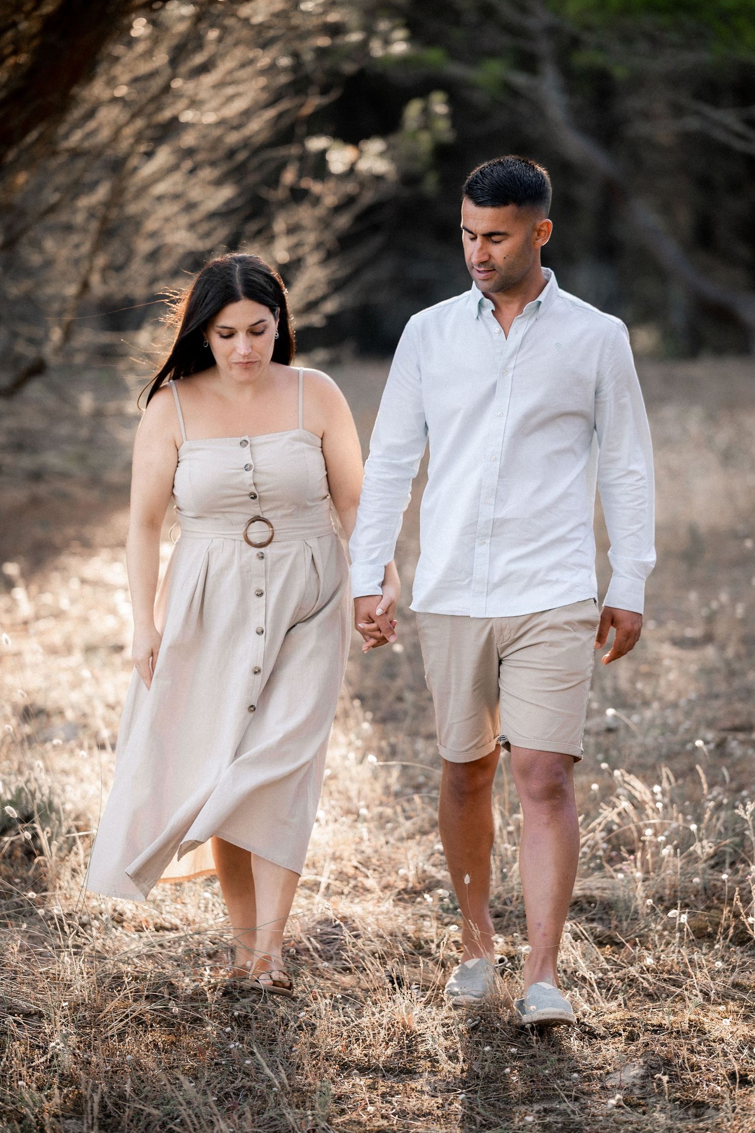 Couple walking together through a sunlit field wearing coordinated beige and white summer outfits.