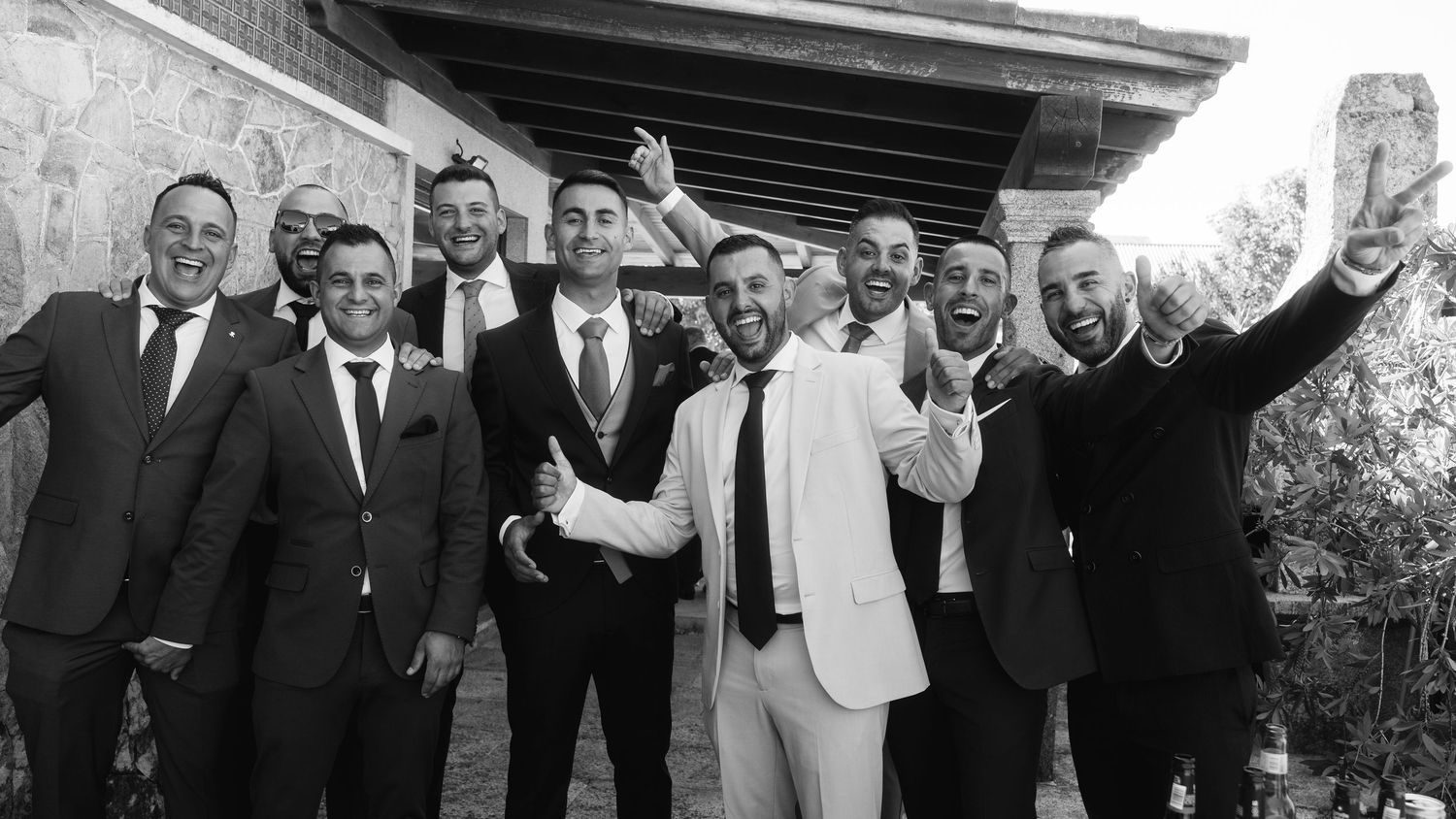 A group of men in formal attire pose together outdoors in black and white, with one person wearing a white jacket.