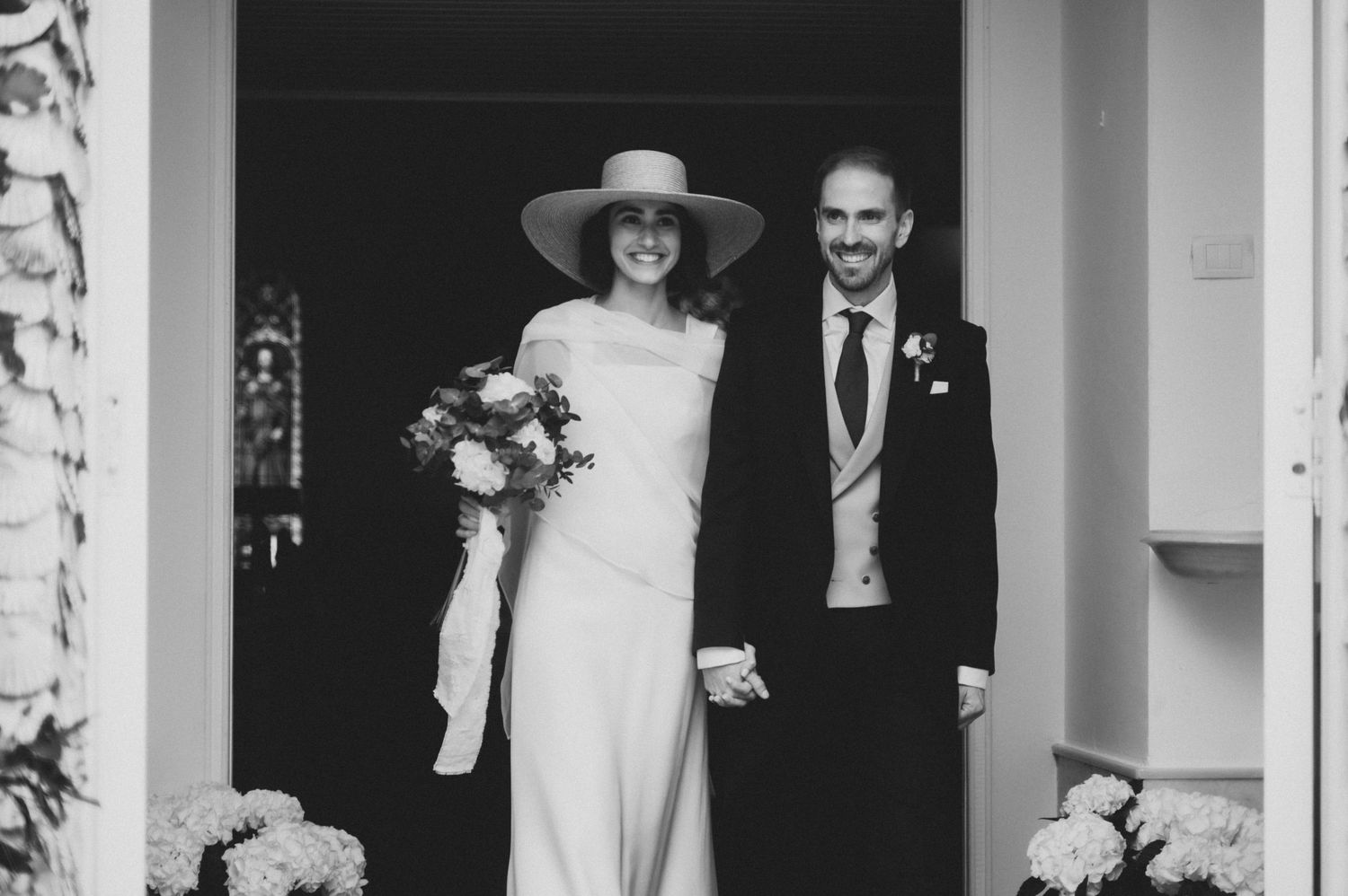 Black and white wedding photo showing a couple walking through an ornate hallway with columns.