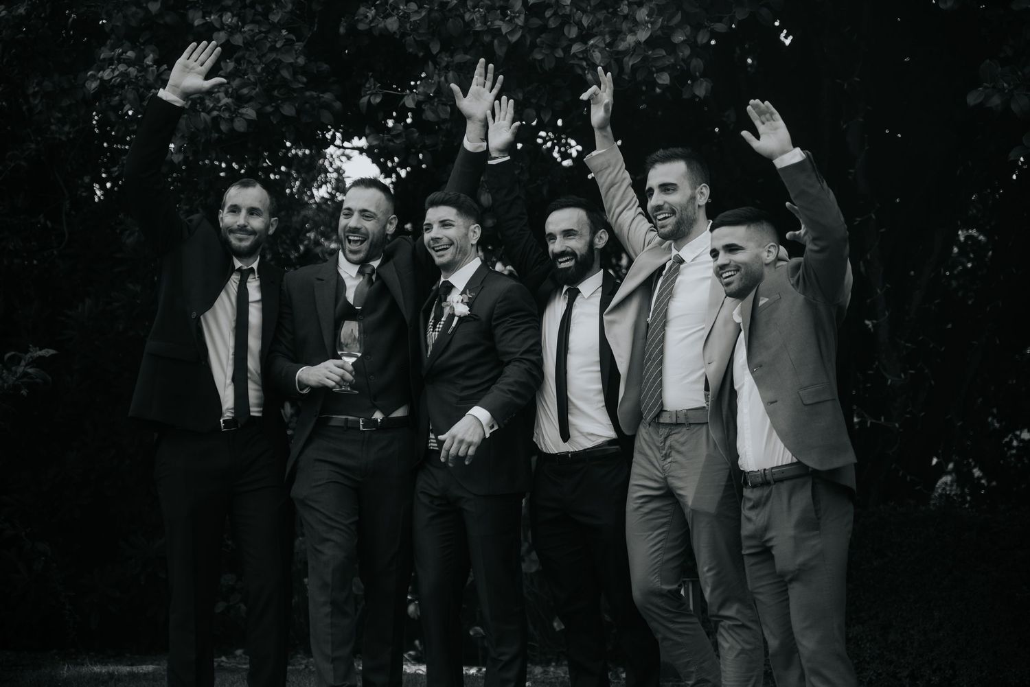 A group of six groomsmen in suits wave their hands in the air while posing together for a black and white photo.