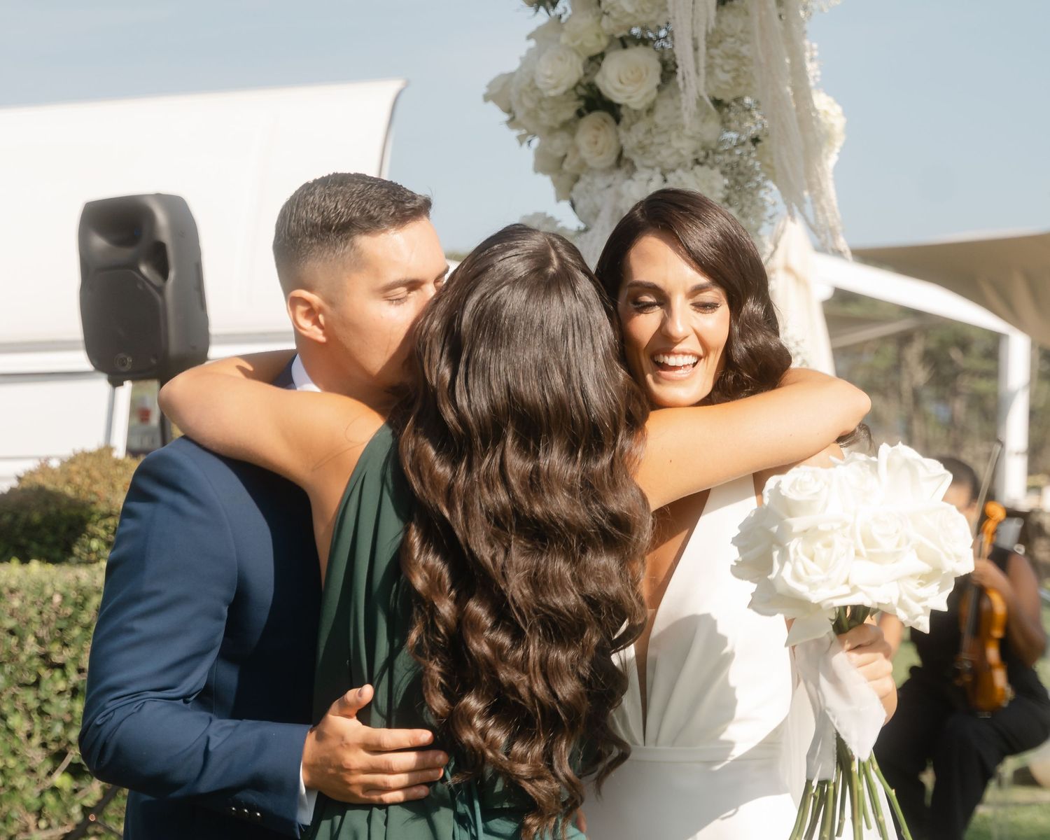 Wedding celebration outdoors with three people embracing while one holds a white floral bouquet.