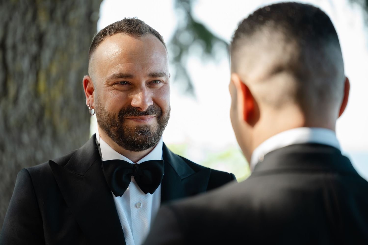 Two people in black tuxedos and bow ties share a tender moment during a wedding ceremony outdoors.