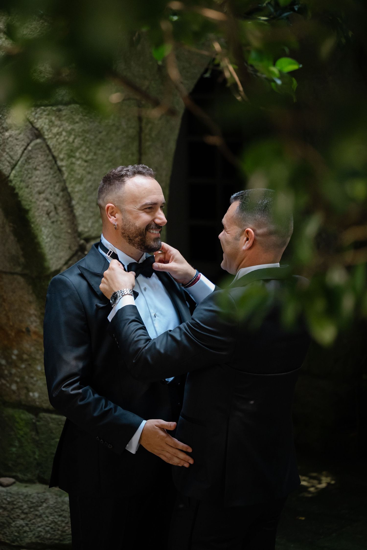 Two grooms in black tuxedos share joyful laughter during an intimate outdoor wedding photoshoot.