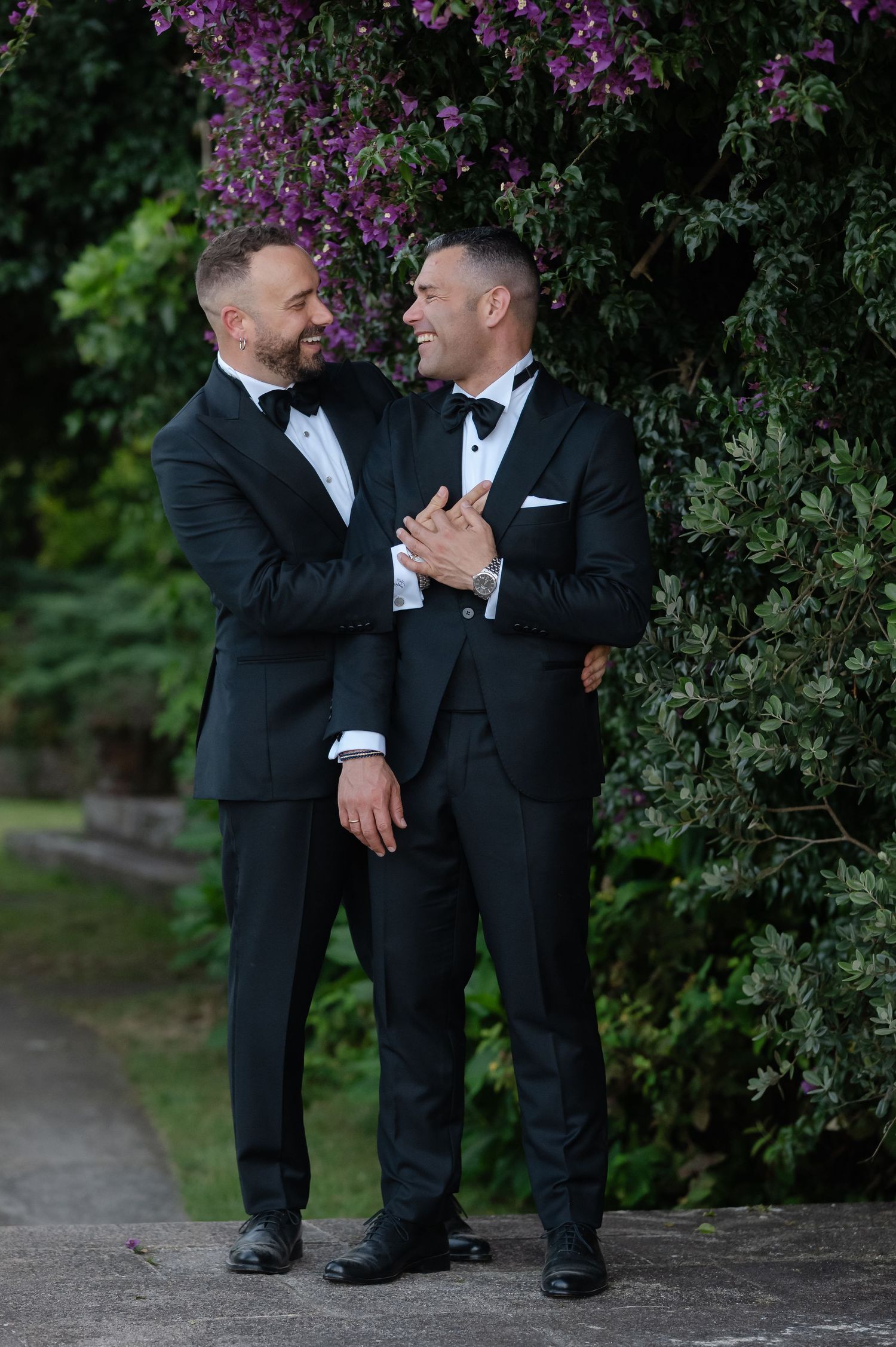 Two people in black suits embrace lovingly under blooming purple flowers in a garden setting.