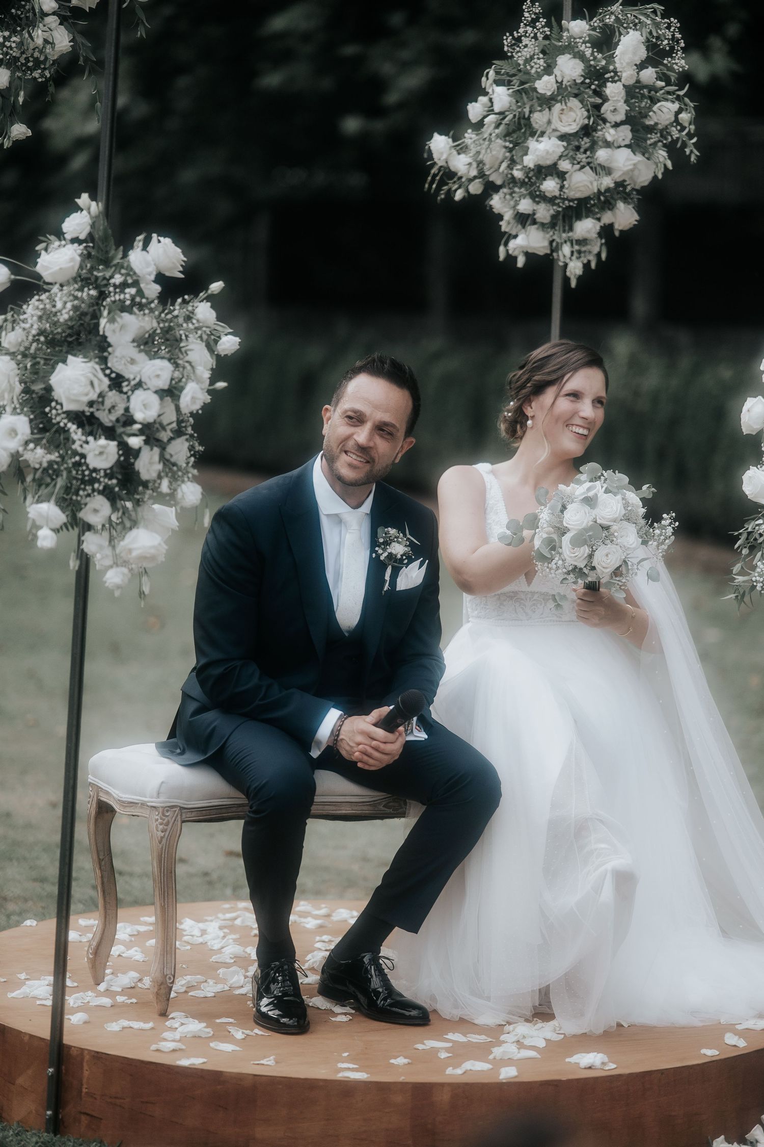 A romantic wedding couple shares a tender moment while sitting on a wooden platform adorned with white floral arrangements.