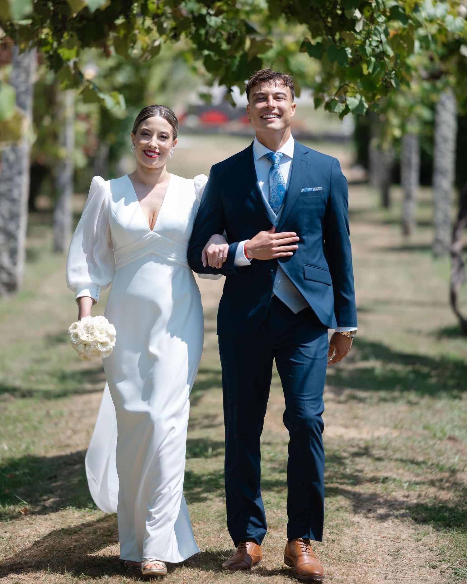 A couple in wedding attire walks together through a sunny garden path lined with trees.