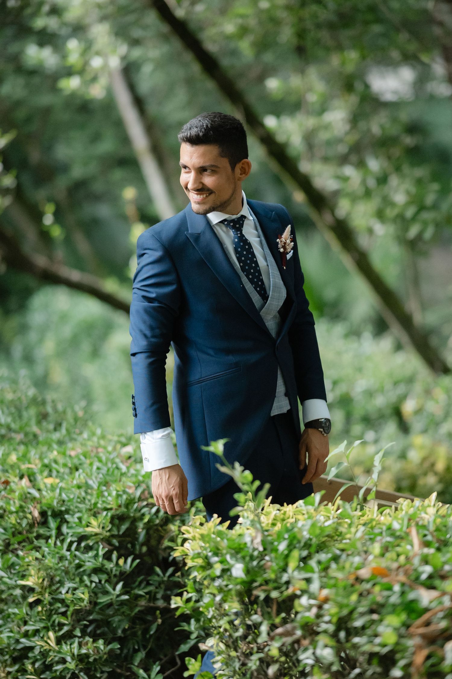 A person in a navy blue suit stands among lush green foliage in an outdoor setting.