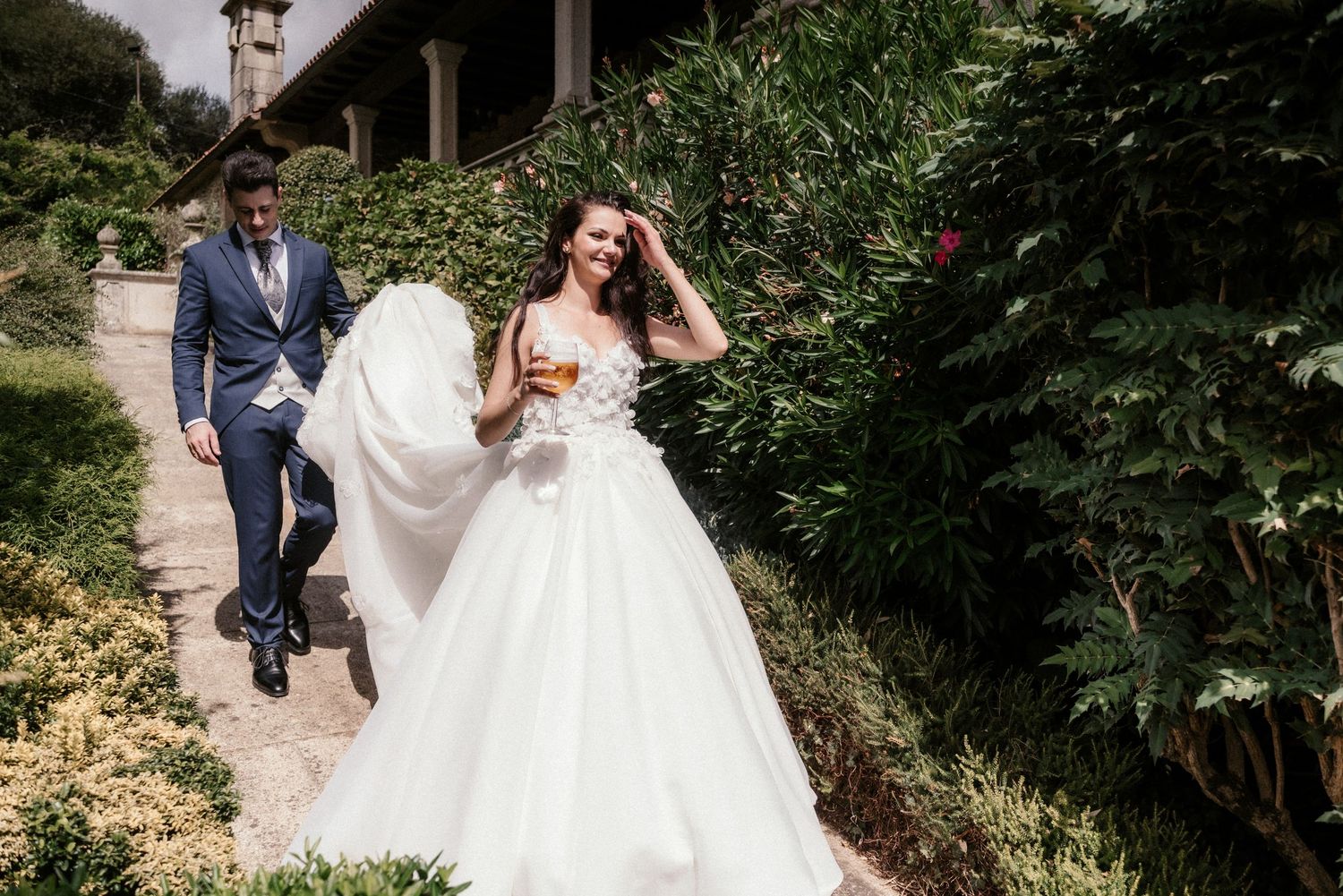 A couple walks along a garden path, with the bride's flowing white dress trailing behind them in an elegant outdoor setting.