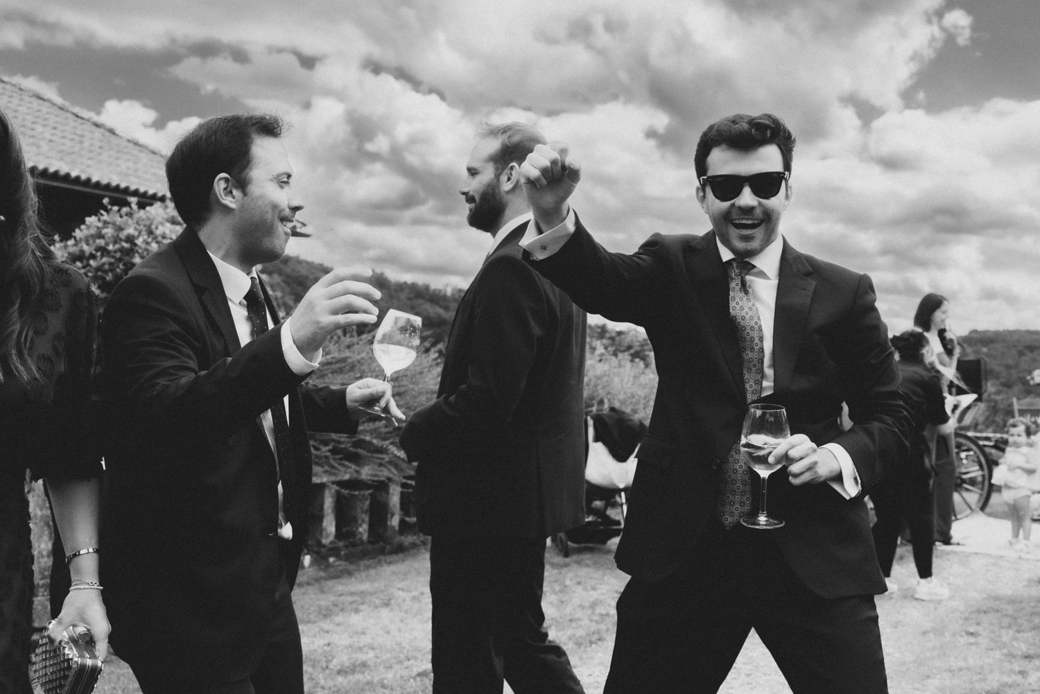 Three men in dark suits and sunglasses share drinks and conversation outdoors on a sunny day with dramatic clouds overhead.