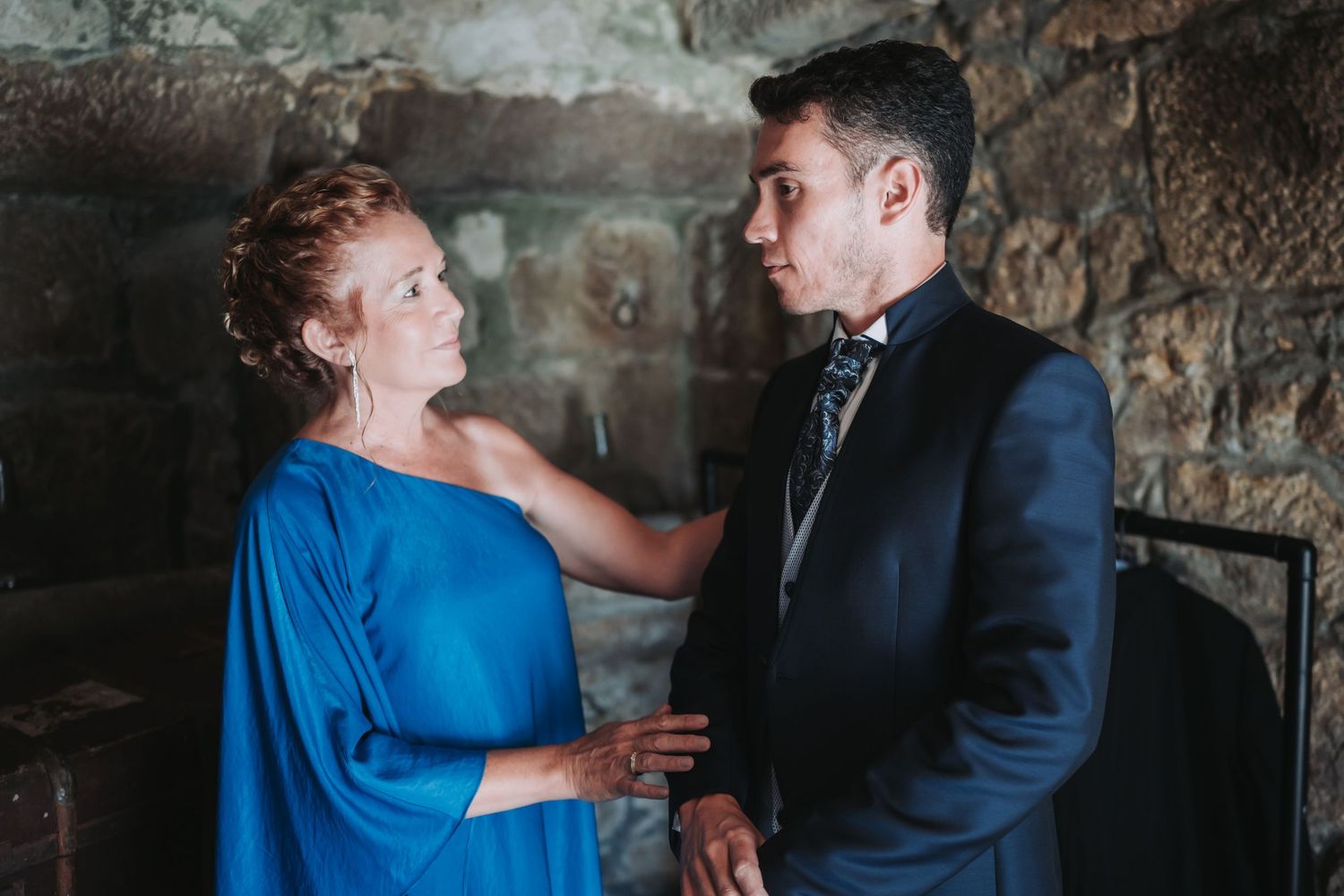 Two people in formal attire share an intimate moment against a rustic stone wall backdrop.