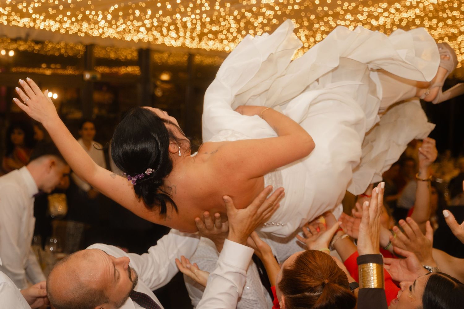 Wedding guests dance and celebrate on a dance floor illuminated by warm golden lights at a reception party.